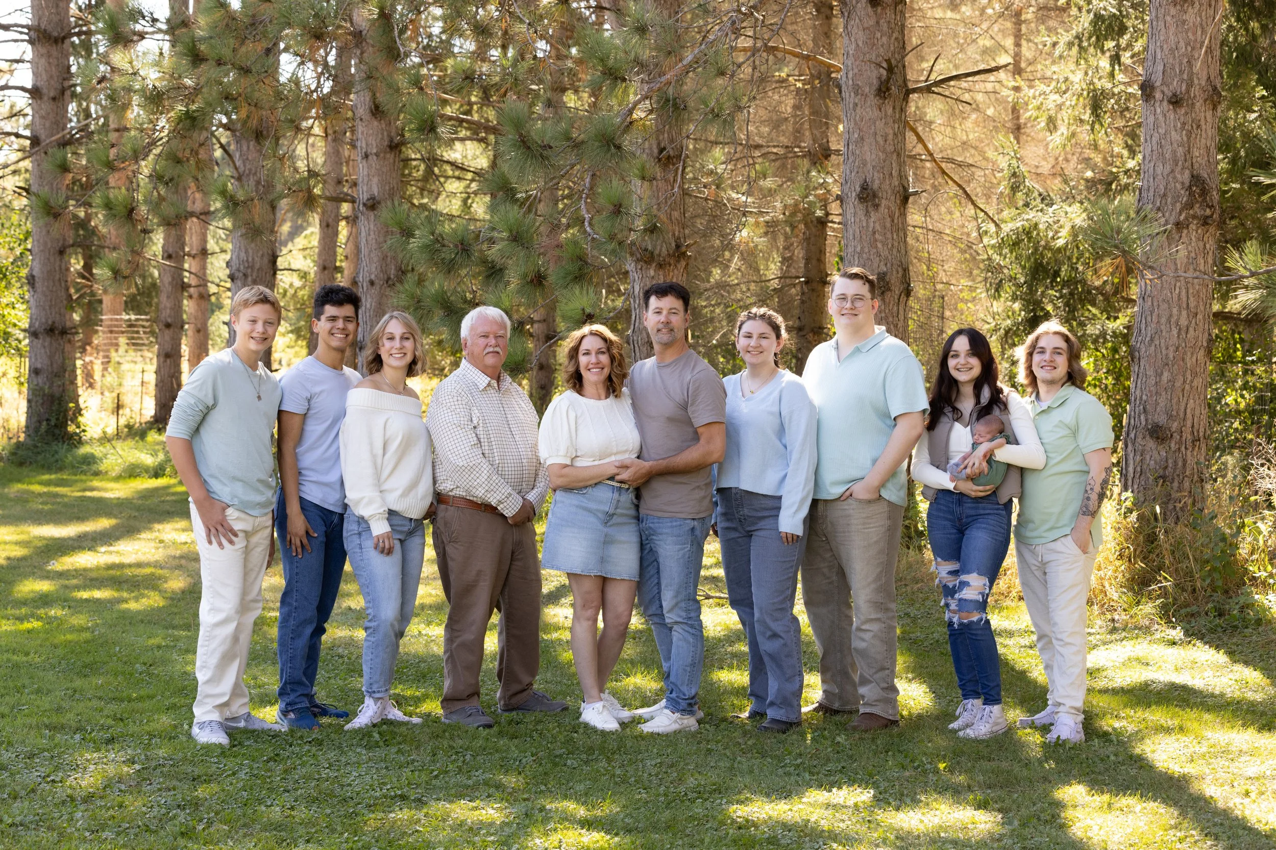 A large family group posing outdoors in a wooded area with tall trees and sunlight filtering through the leaves. The family members are standing in a line, smiling at the camera.