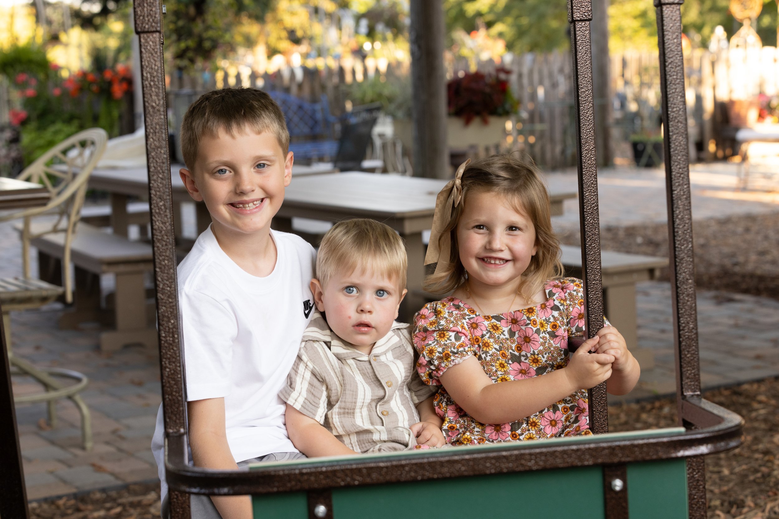 Children sitting on a park or playground swing, smiling and enjoying outdoors in a backyard setting with trees and wooden furniture.