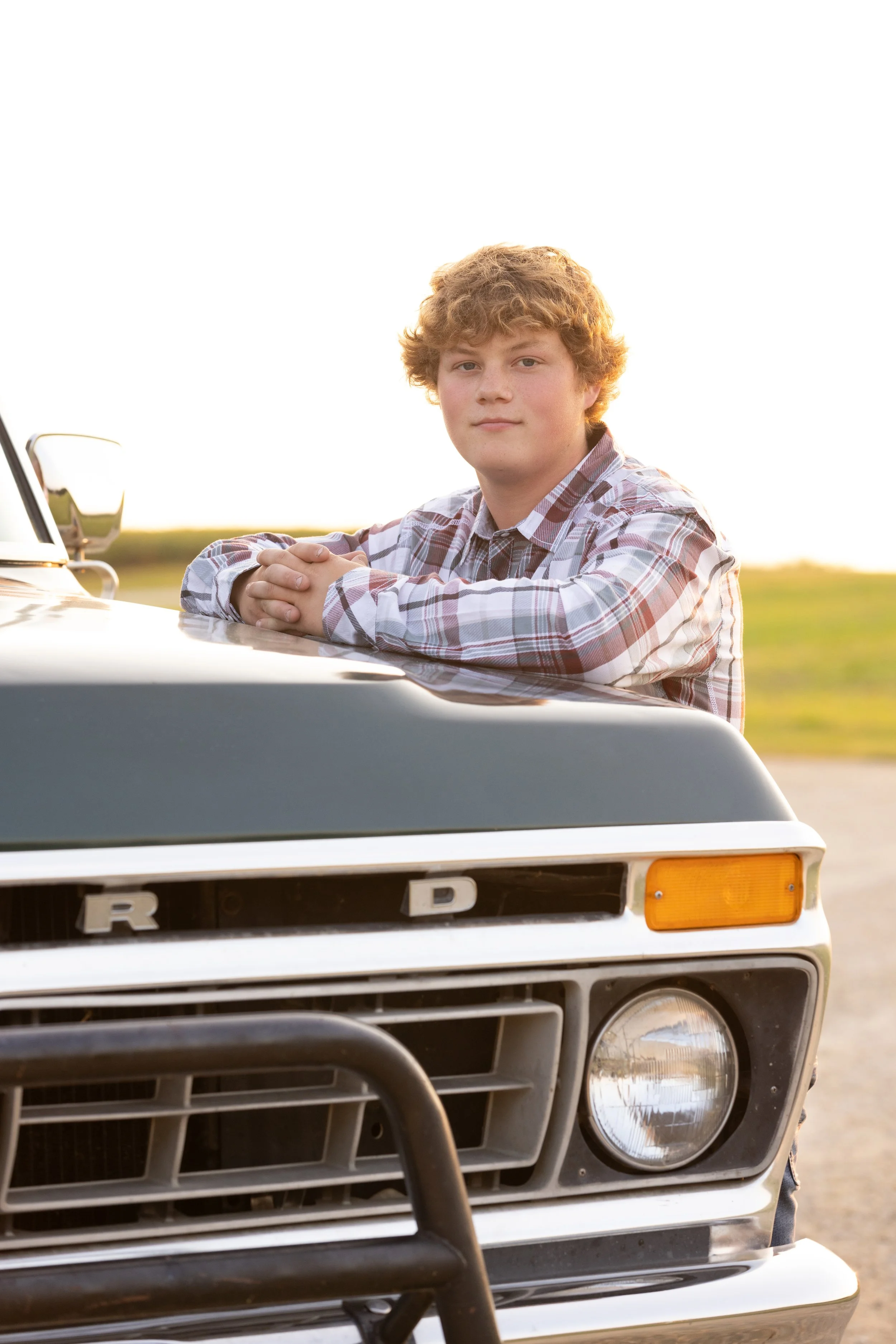 A young man with curly blond hair leaning on the hood of a black vintage Ford truck outdoors during sunset or sunrise, wearing a plaid shirt.