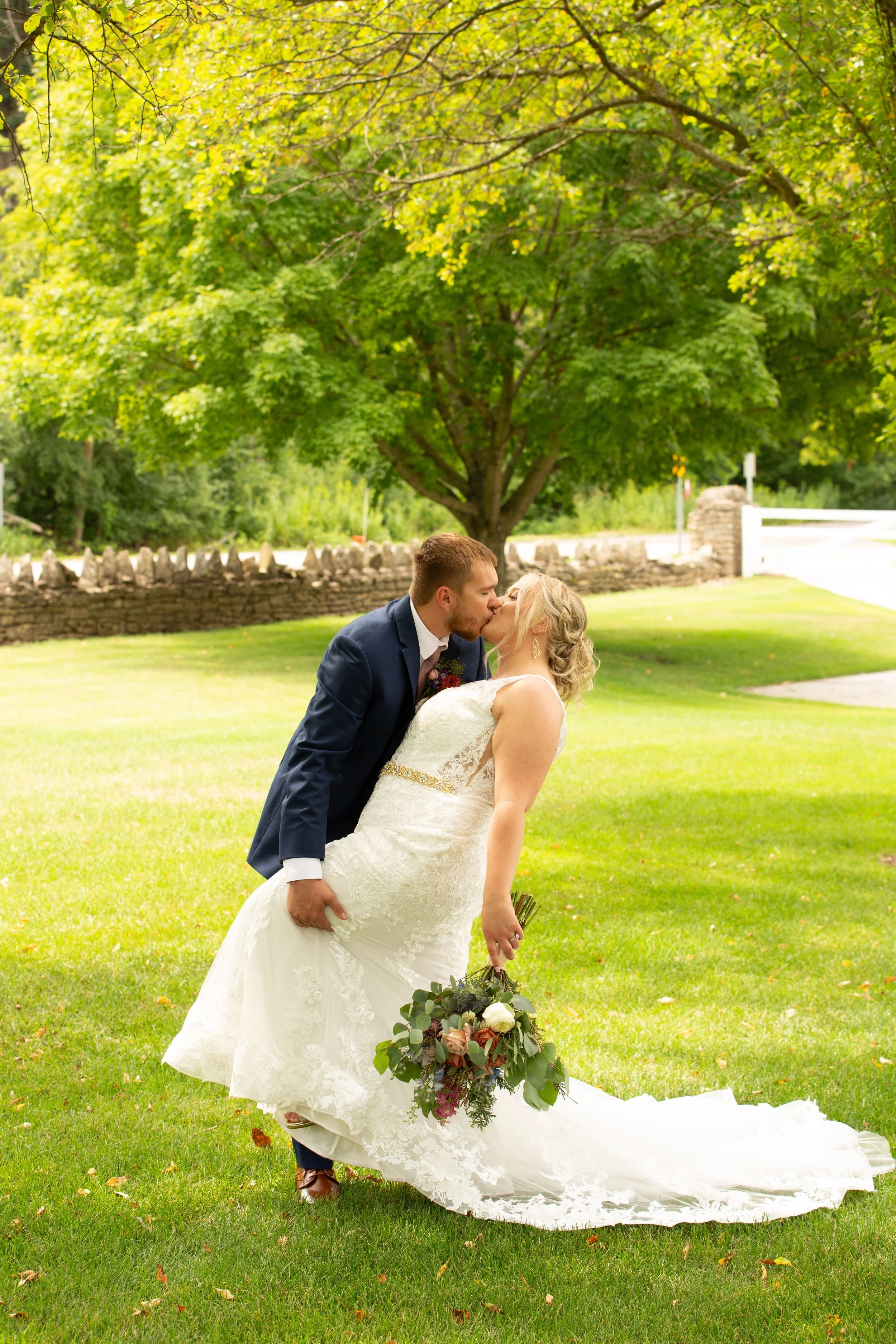 A newlywed couple sharing a kiss outdoors on a green lawn with trees in the background. The groom is wearing a navy blue suit, and the bride is in a white wedding gown holding a bouquet of flowers.
