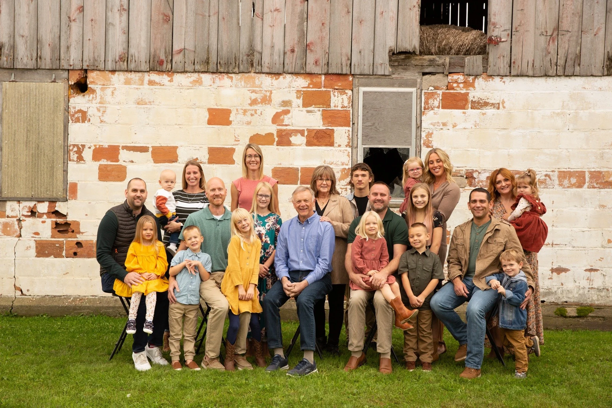 A large family gathering outside in front of a rustic barn with peeling paint and weathered wood, featuring adults and children smiling and sitting or standing on green grass.