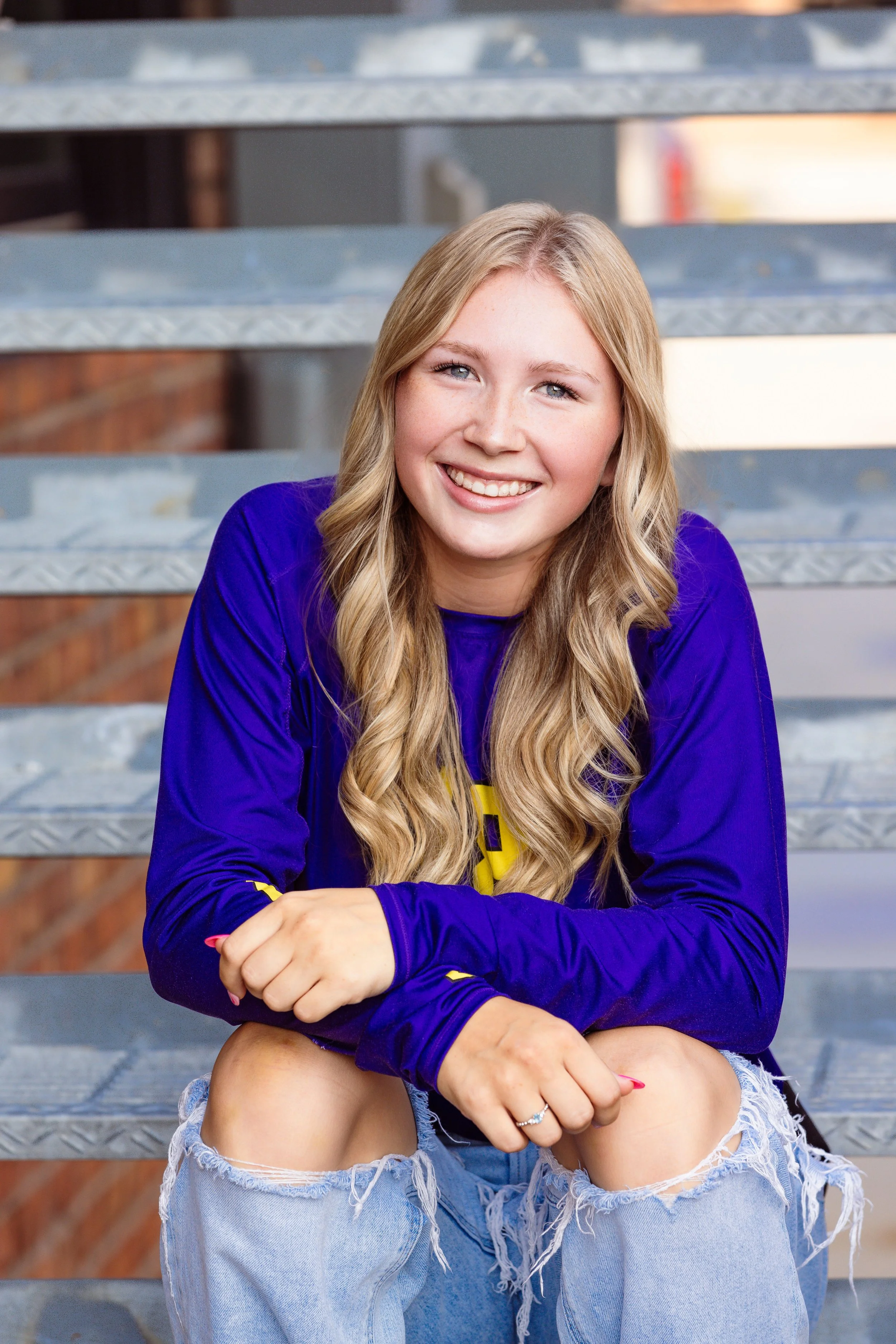 A young woman sitting on metal bleachers, smiling, wearing a purple long sleeve shirt and ripped jeans.