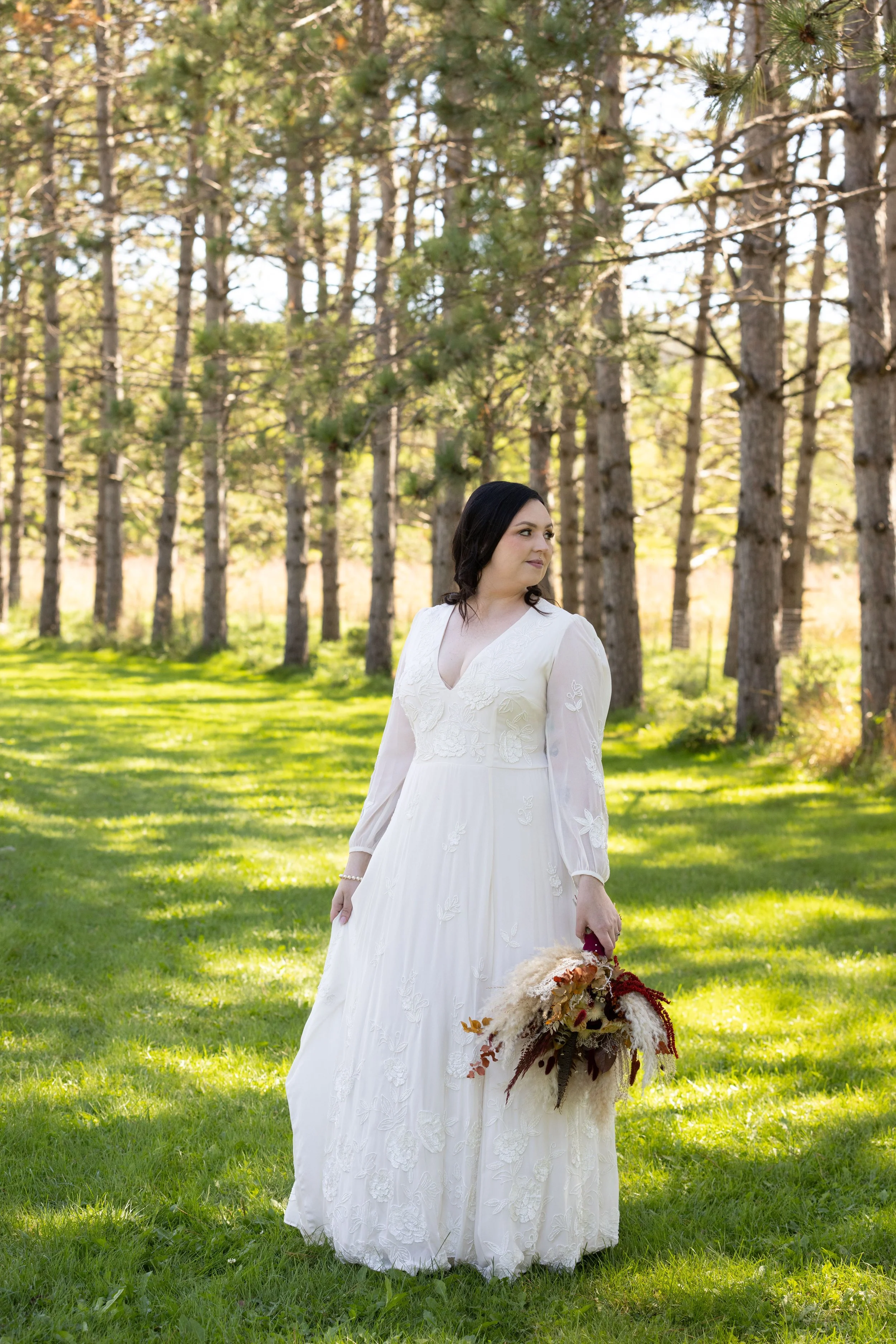 A woman in a white wedding dress holding a bouquet of flowers standing in a wooded grassy area.