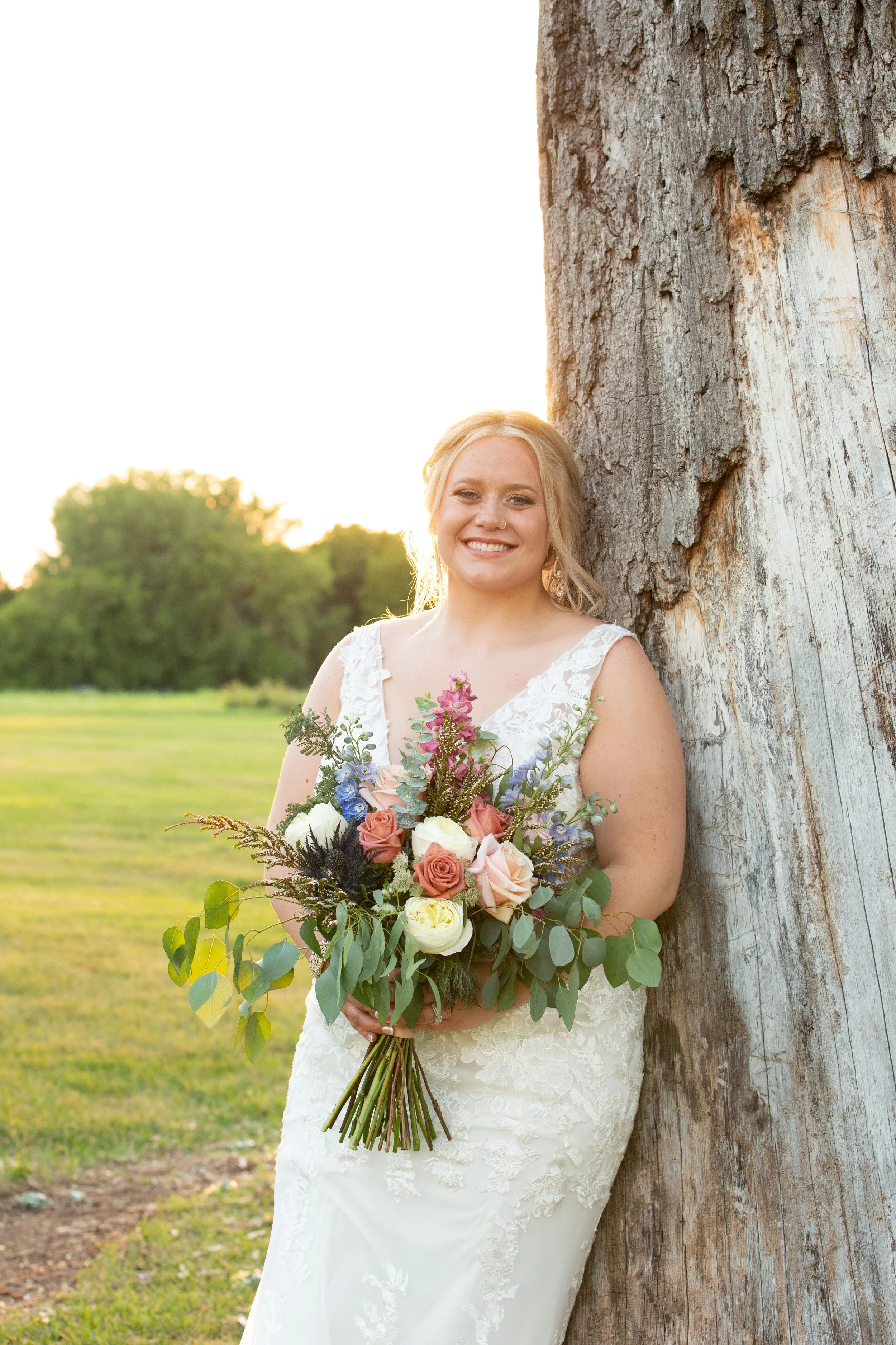 A woman in a white lace wedding dress holding a colorful bouquet of roses and greenery, standing outdoors next to a large tree at sunset.