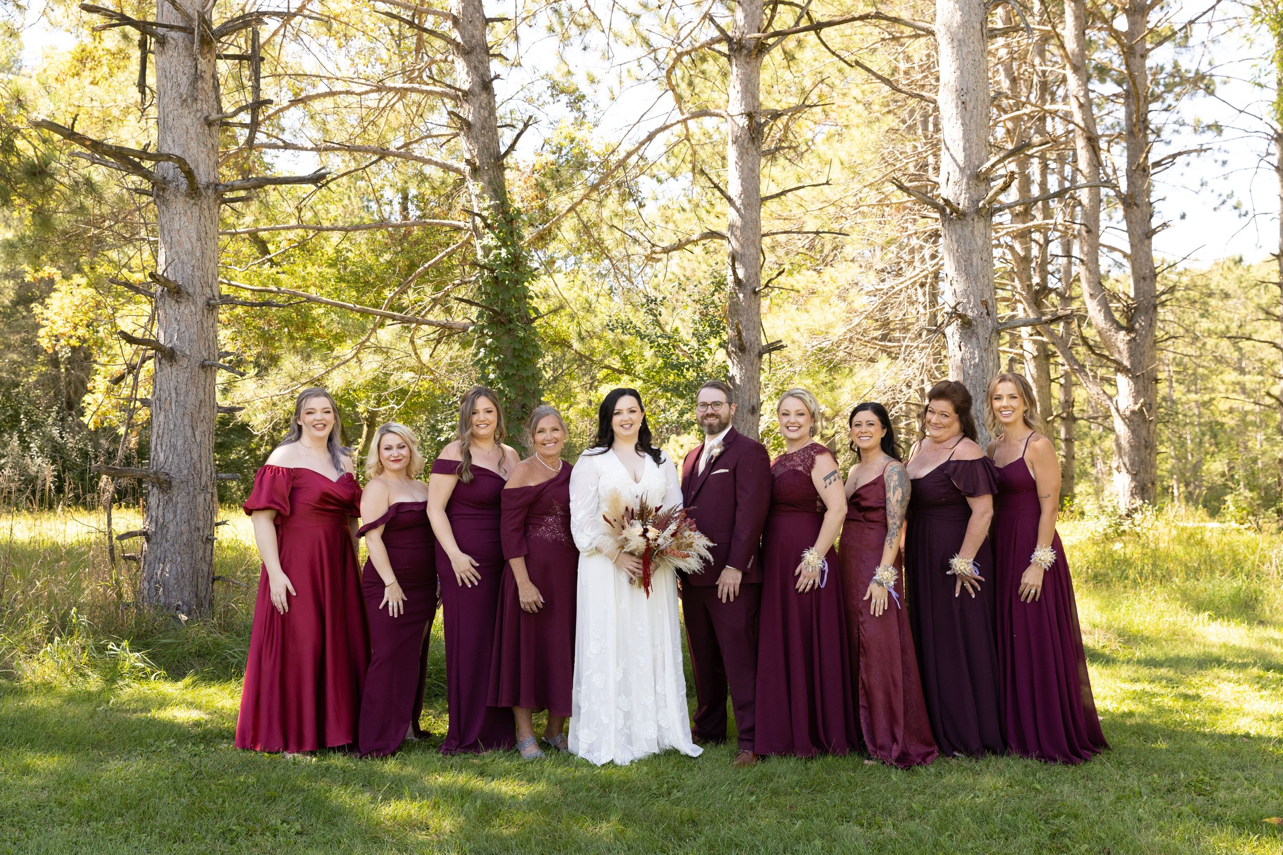 A wedding party with bridesmaids and a bride and groom standing outdoors in a forested area during daytime.