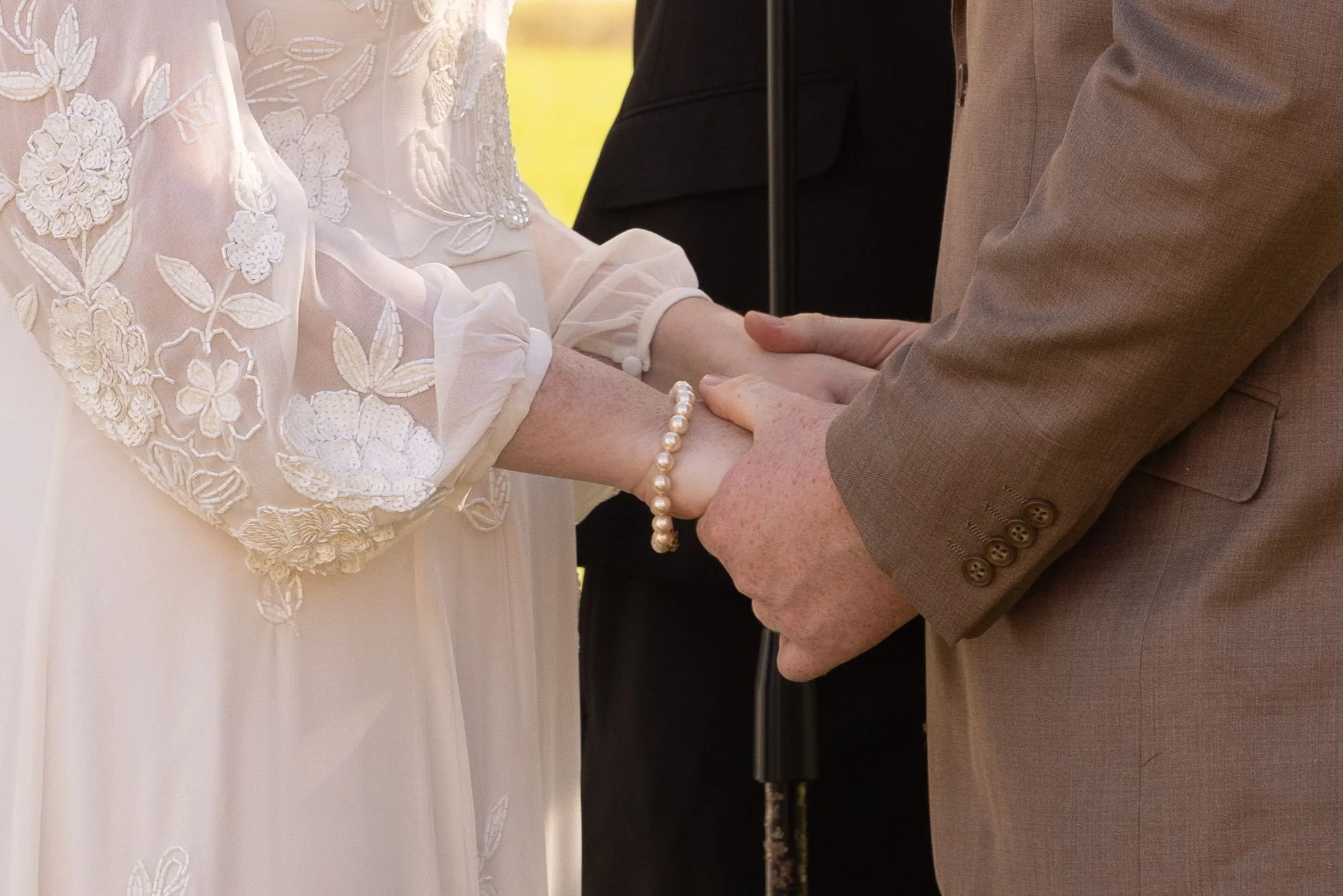 A bride and groom holding hands during a wedding ceremony, with the bride wearing a lace-embellished wedding dress and a pearl bracelet, and the groom in a light brown suit.