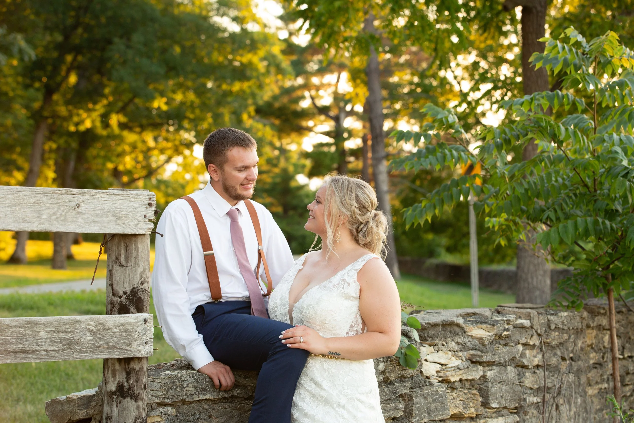 A couple dressed in wedding attire sitting outdoors on a stone wall, smiling at each other during sunset in a park with green trees.