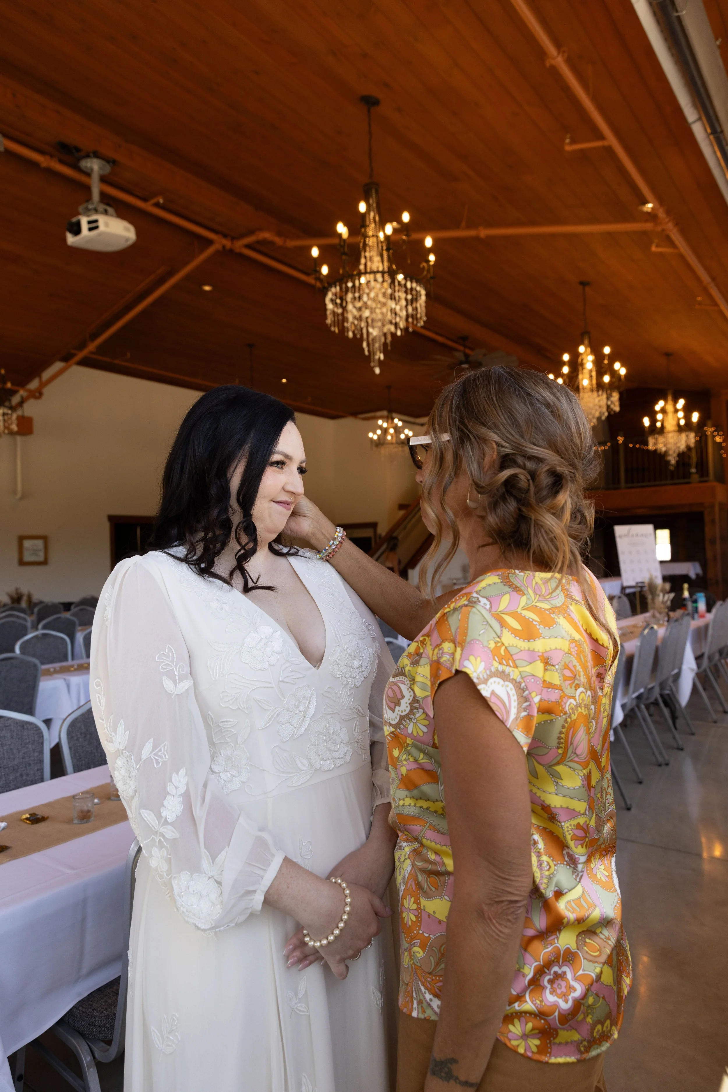 A woman in a white dress faces another woman in a colorful floral shirt. The woman in white is smiling softly, and their foreheads touch in an intimate moment inside a decorated venue with chandeliers and tables.