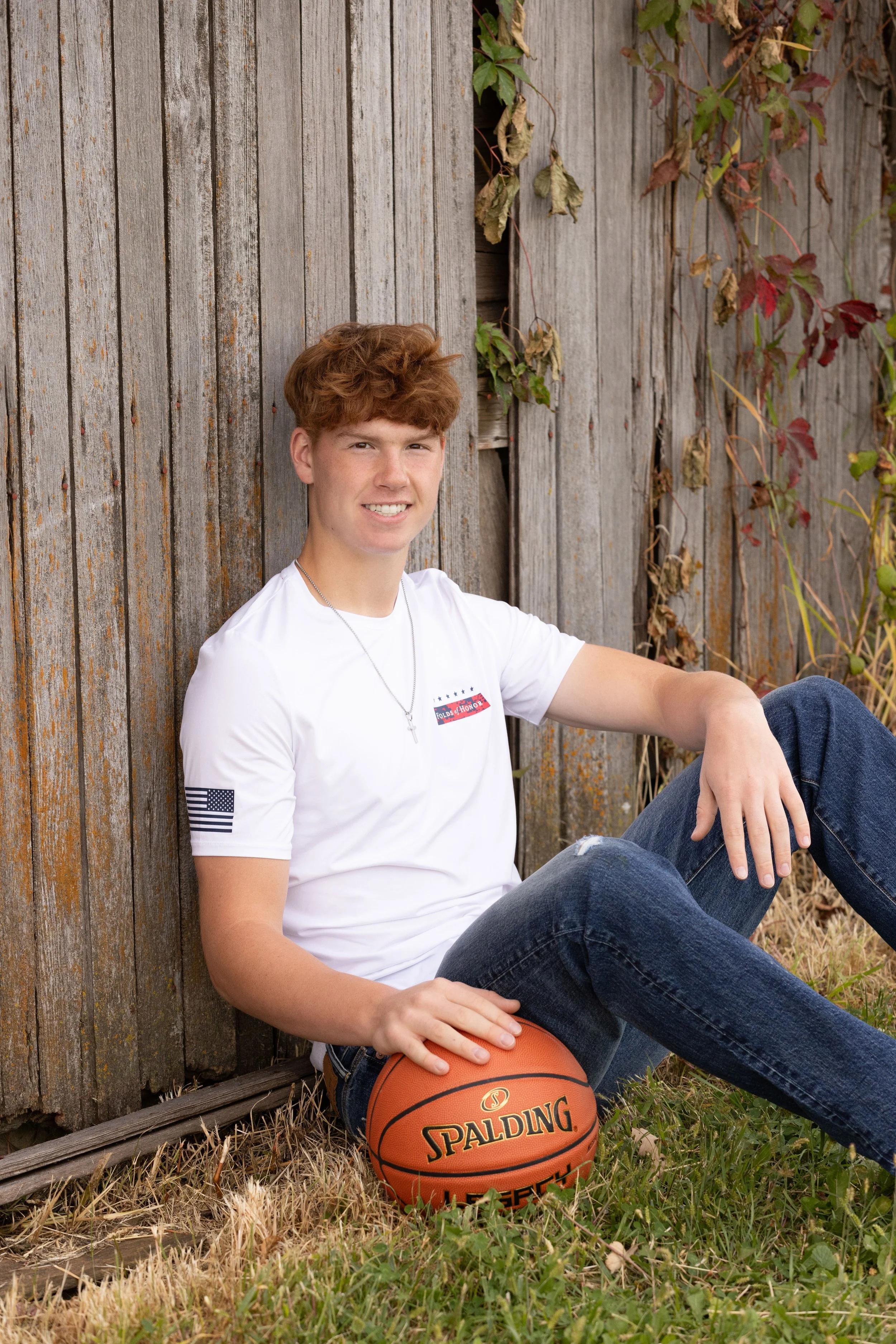A young man with red hair sitting on the grass next to a weathered wooden fence, holding an orange Spalding basketball, smiling at the camera, wearing a white T-shirt with American flag accents and blue jeans.