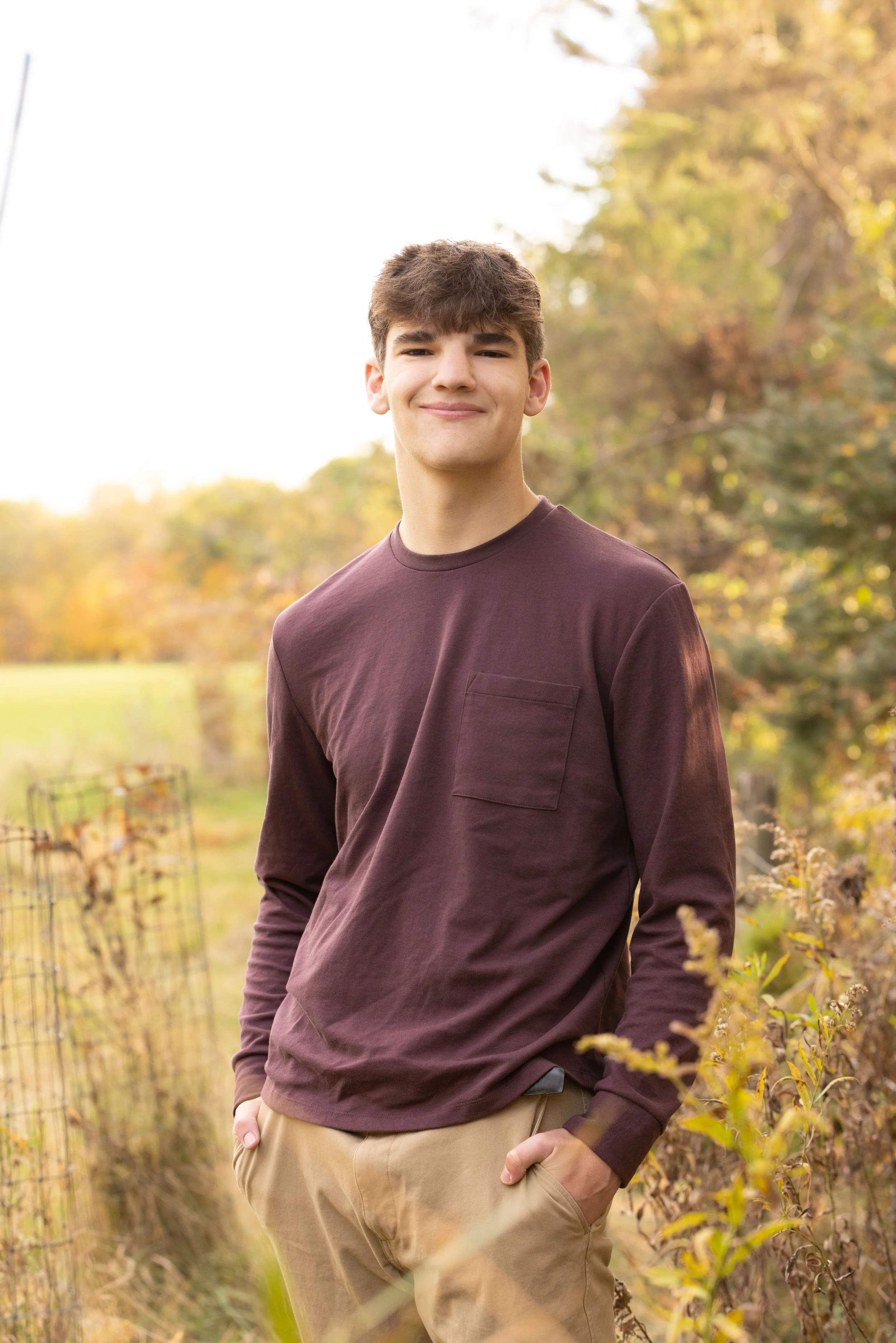 A young man with brown hair smiling outdoors in a rural, autumn setting. He is wearing a maroon long-sleeve shirt and beige pants, with hands in pockets, standing beside a wire fence among golden foliage.