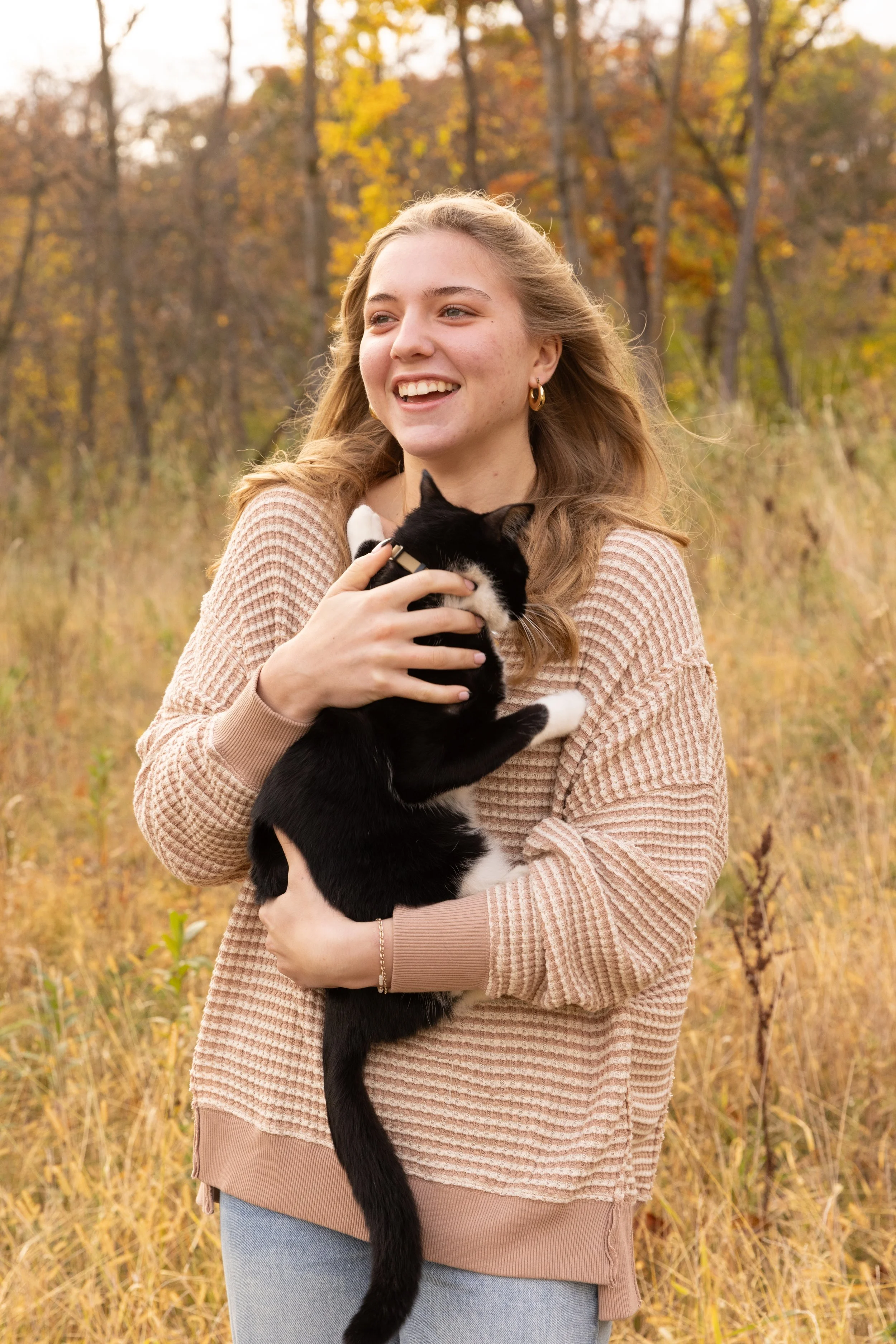 A young woman with long wavy hair, wearing a beige knit sweater and jeans, is smiling while holding a black and white cat in an outdoor setting with fall foliage in the background.