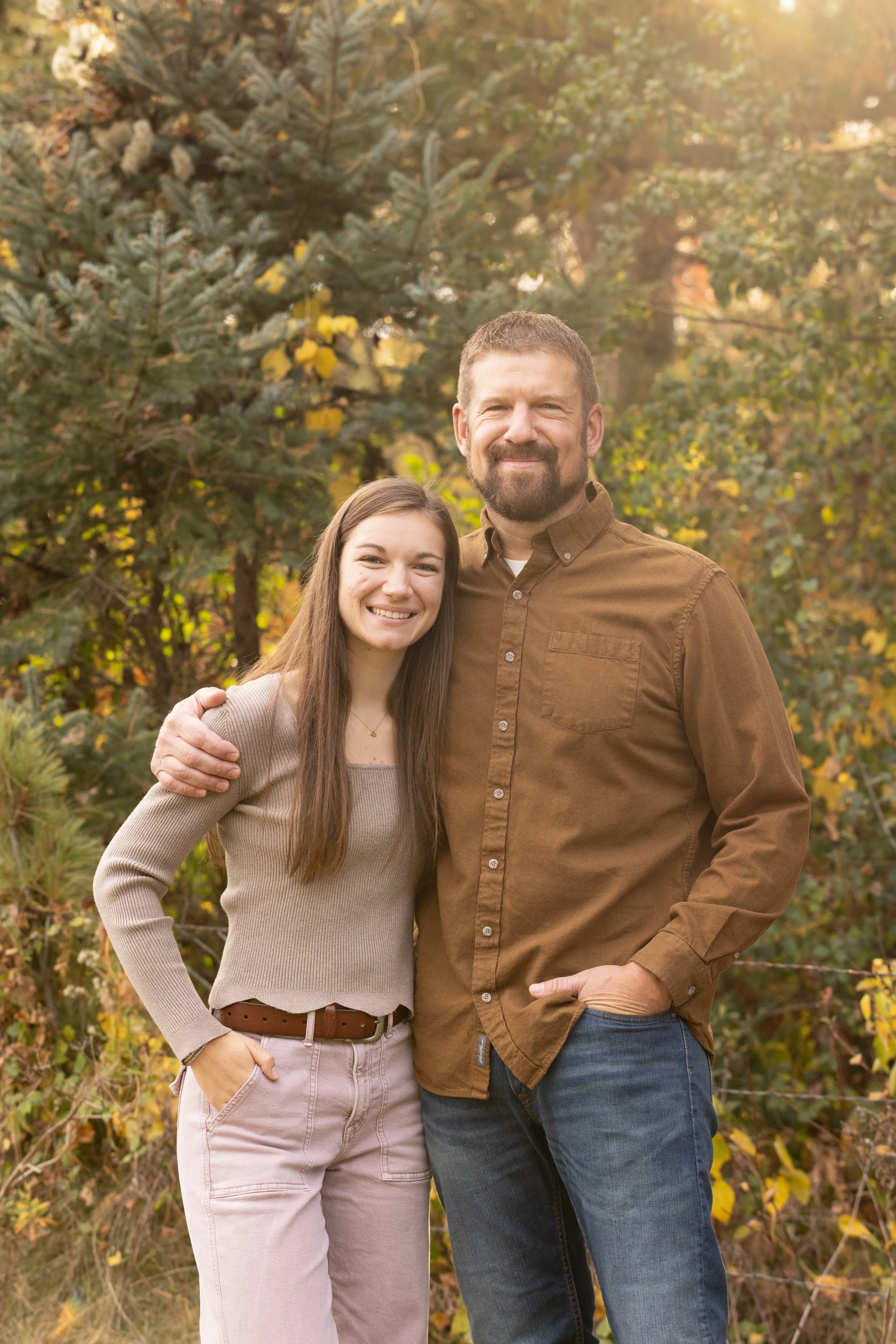 A young woman and an older man stand together outdoors in front of trees, smiling warmly at the camera during sunset.