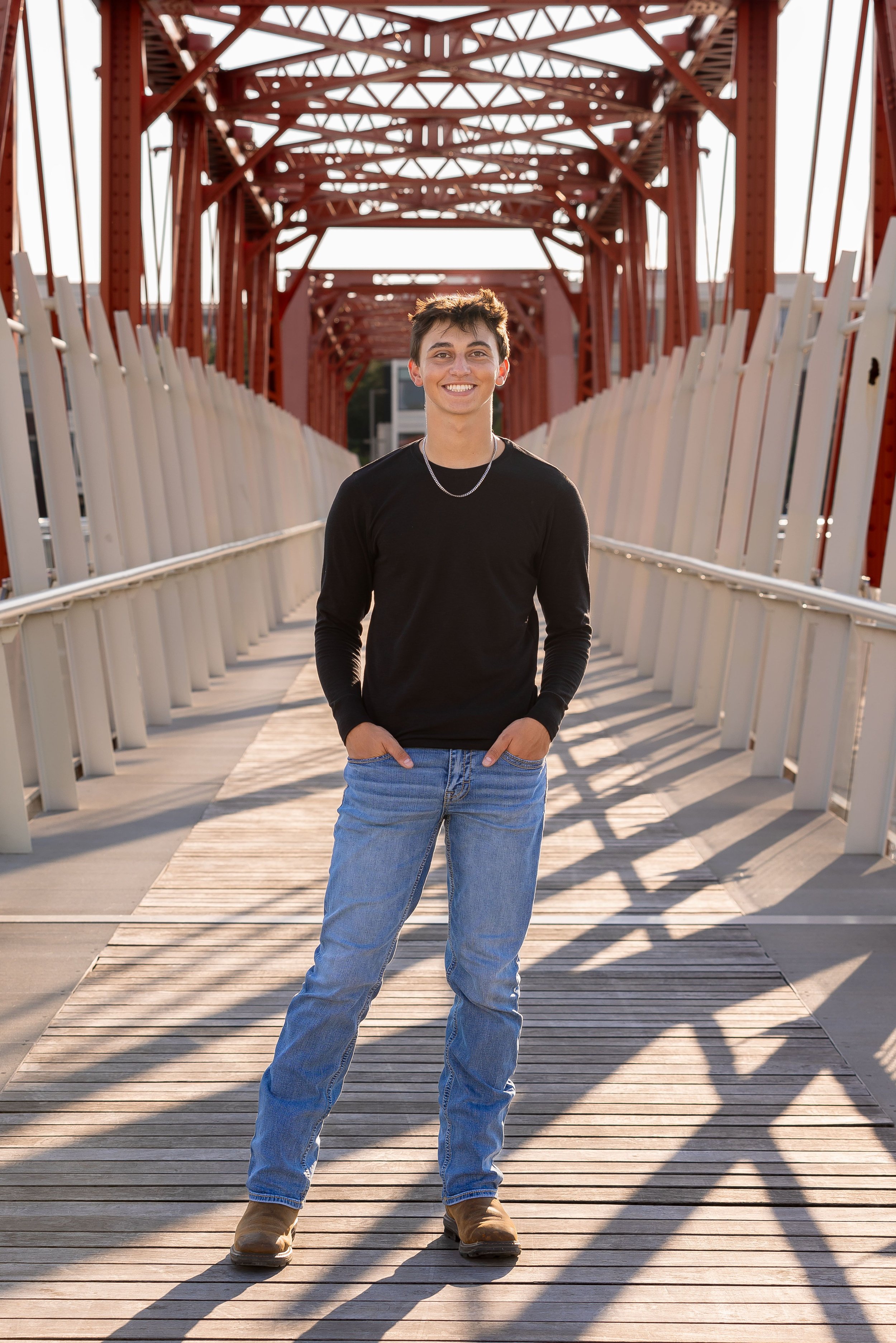 A young man smiling and standing with hands in pockets on a pedestrian bridge with red metal framework and wooden flooring.
