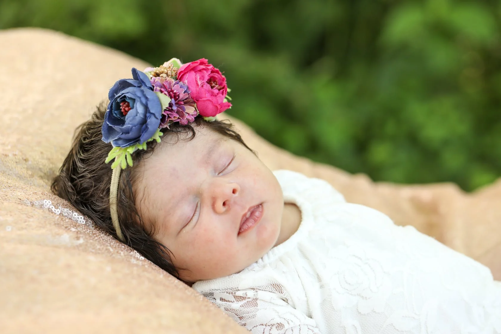 A sleeping baby girl with dark brown hair, lying on a light brown surface, wearing a white dress and a colorful floral headband with pink and blue flowers, with blurred green foliage in the background.