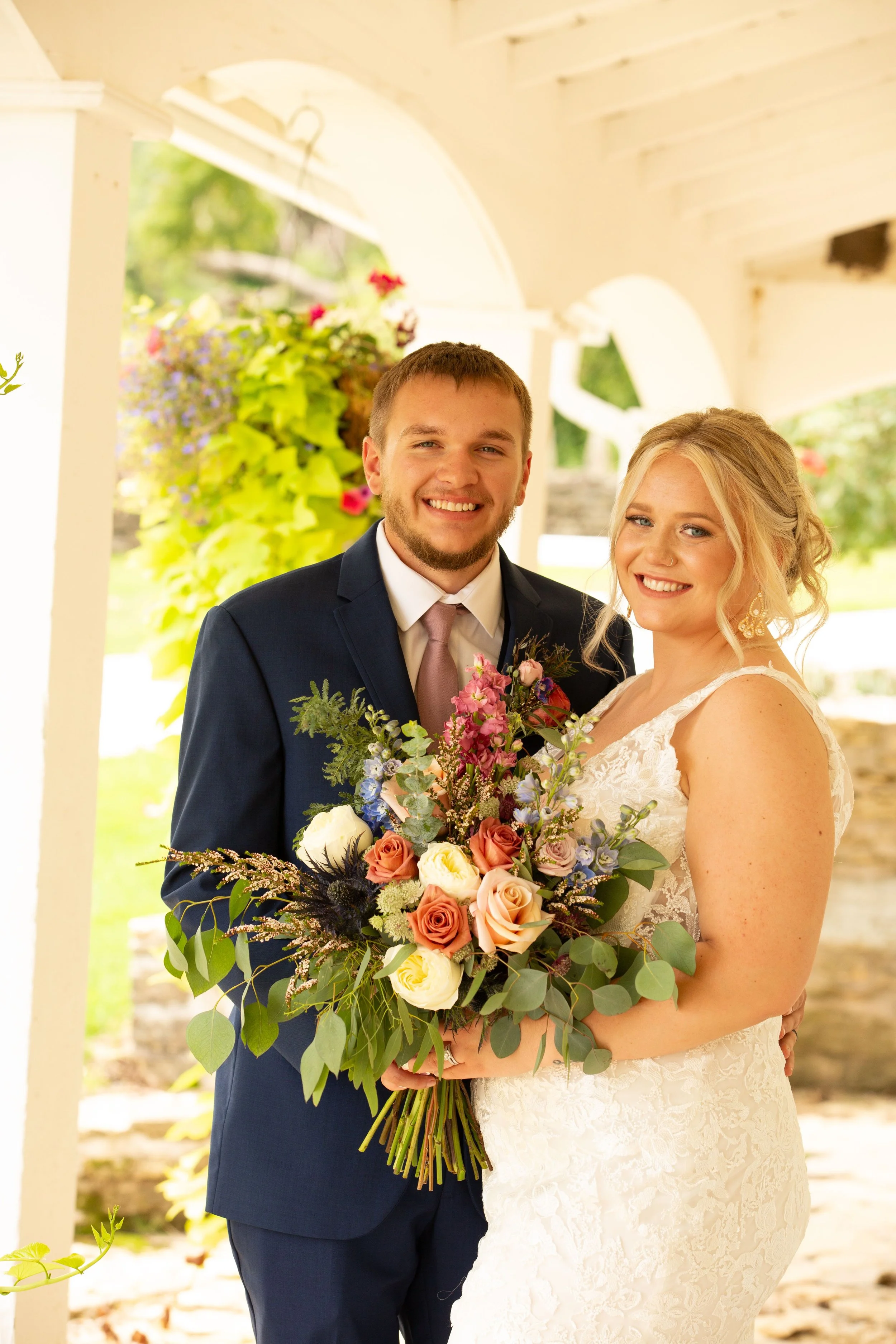 A newlywed couple stands under a porch, smiling at the camera, with the bride holding a large bouquet of flowers.