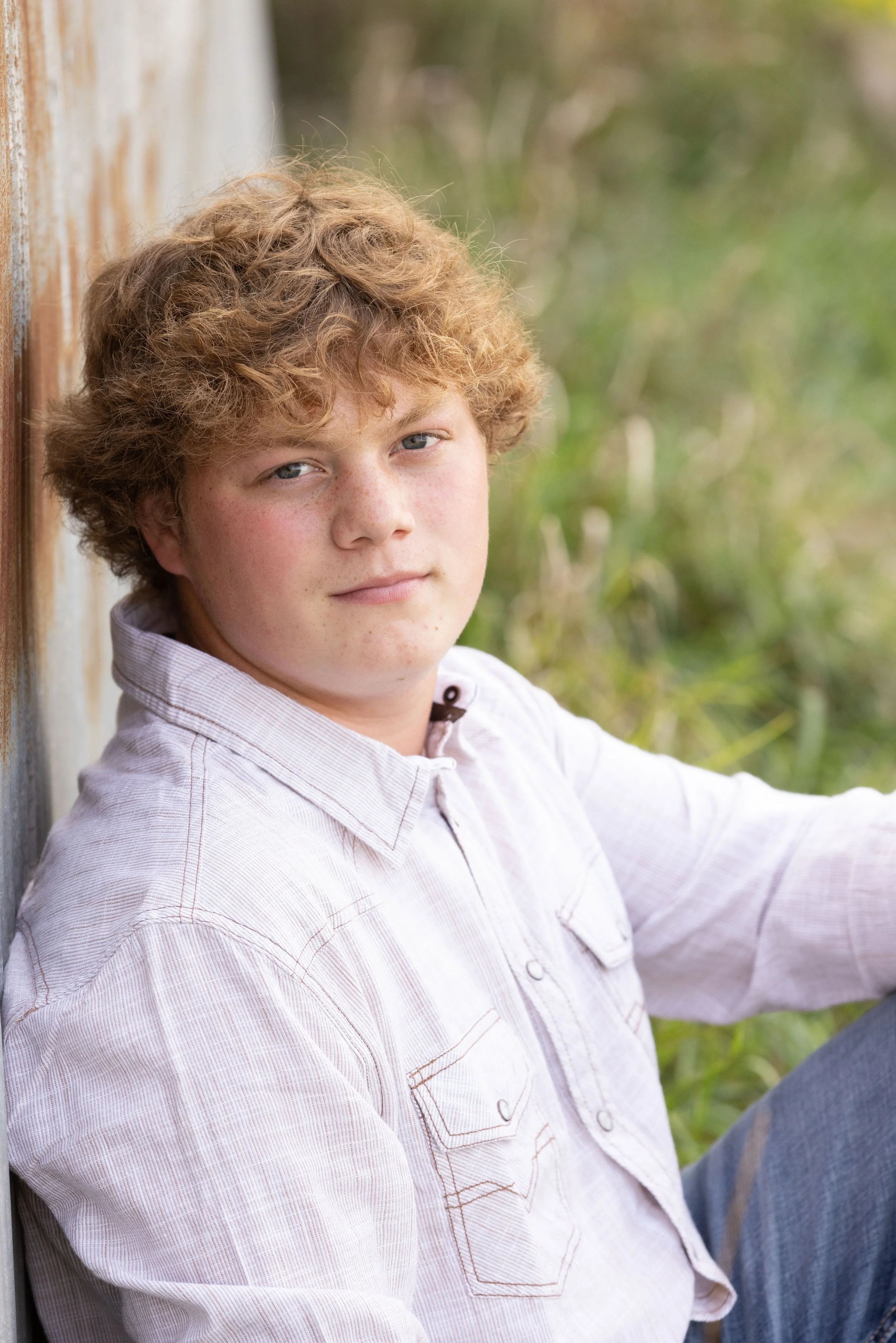 A young boy with curly blond hair and light skin, wearing a light-colored button-up shirt, sitting outdoors against a weathered wooden wall, with blurred green foliage in the background.
