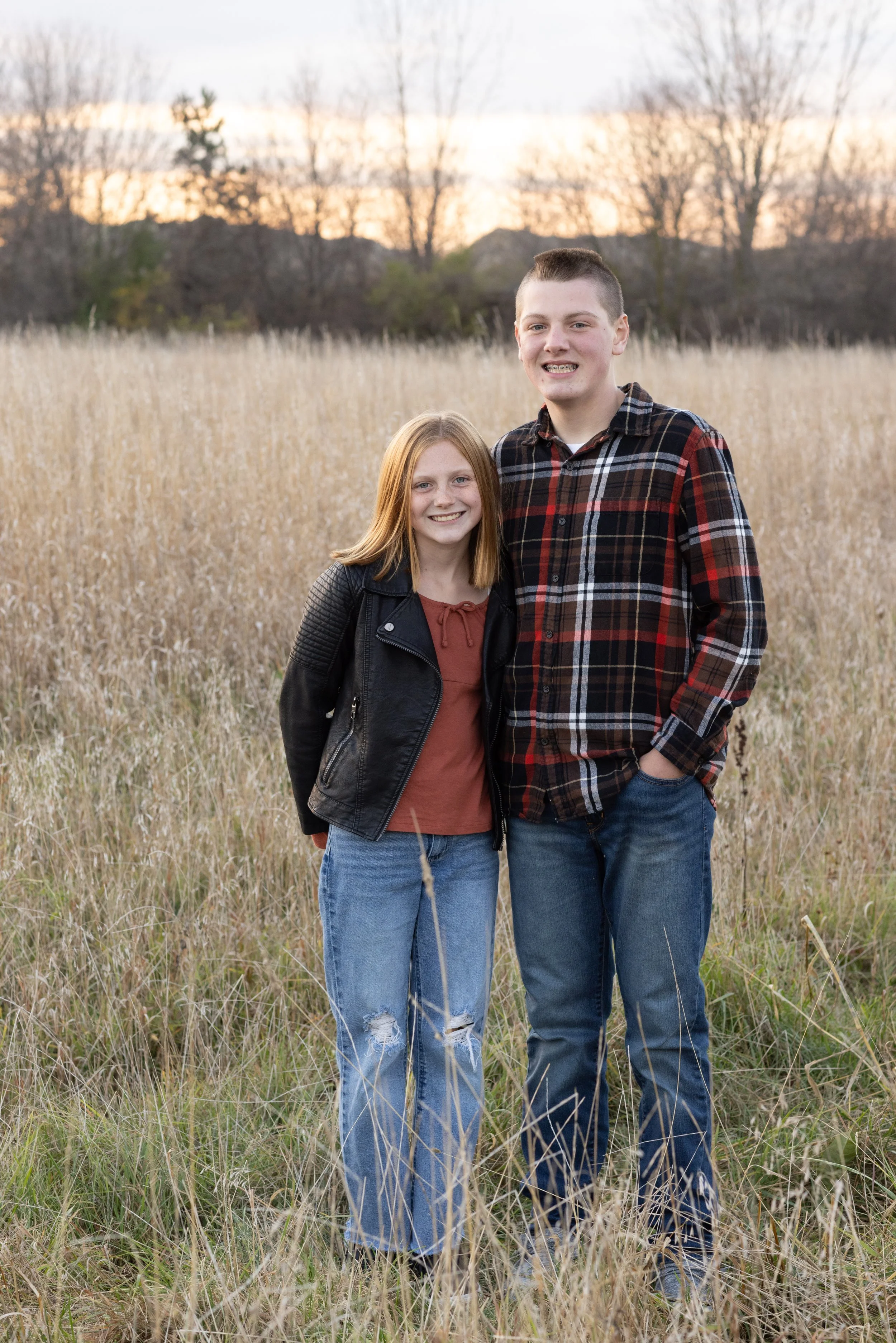 A teenage girl and teenage boy standing together in a field of tall grass, smiling at the camera during sunset.
