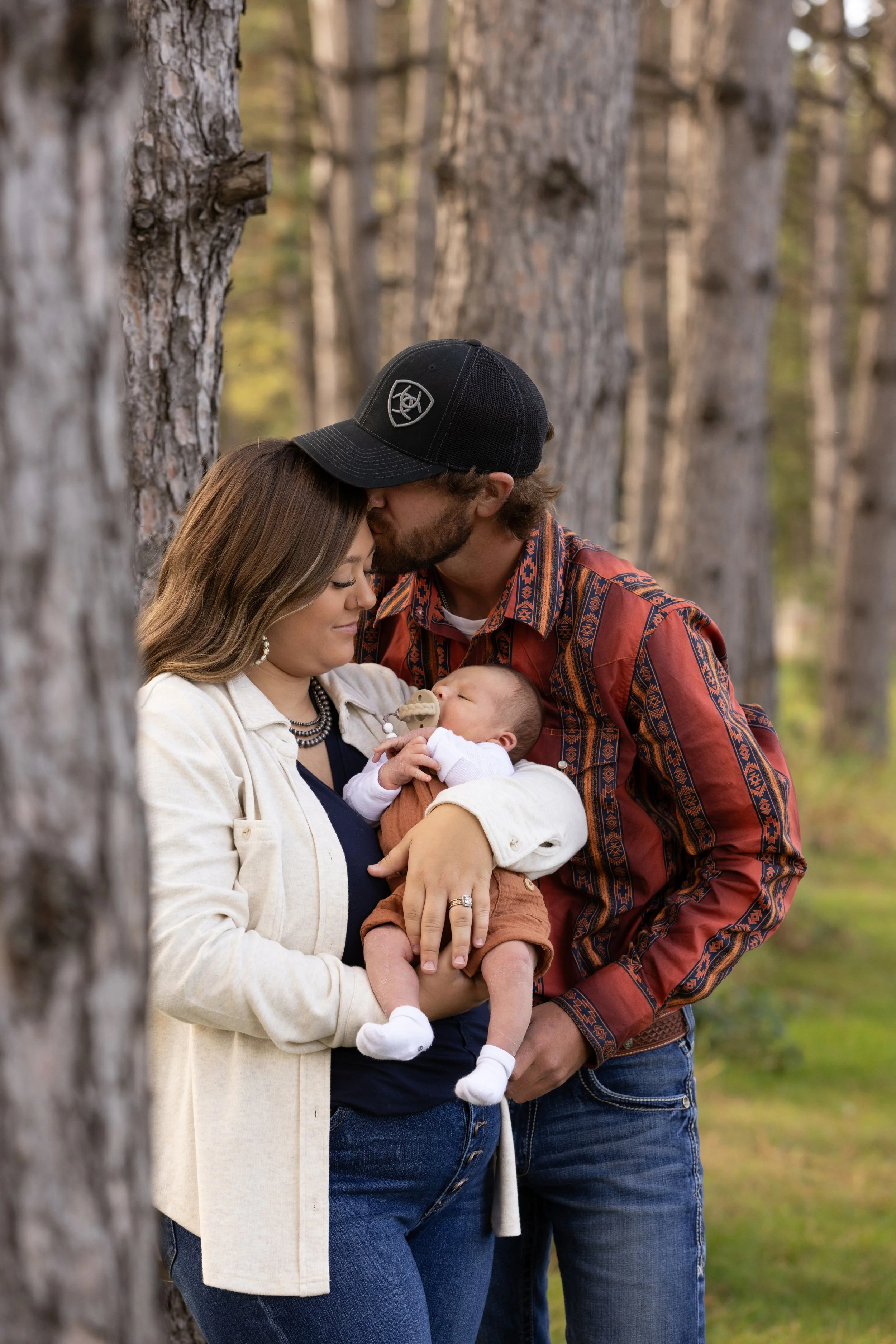 A family of three with a newborn, standing outdoors among tall trees, sharing a tender moment as the father gently kisses the mother's forehead while she holds the baby.