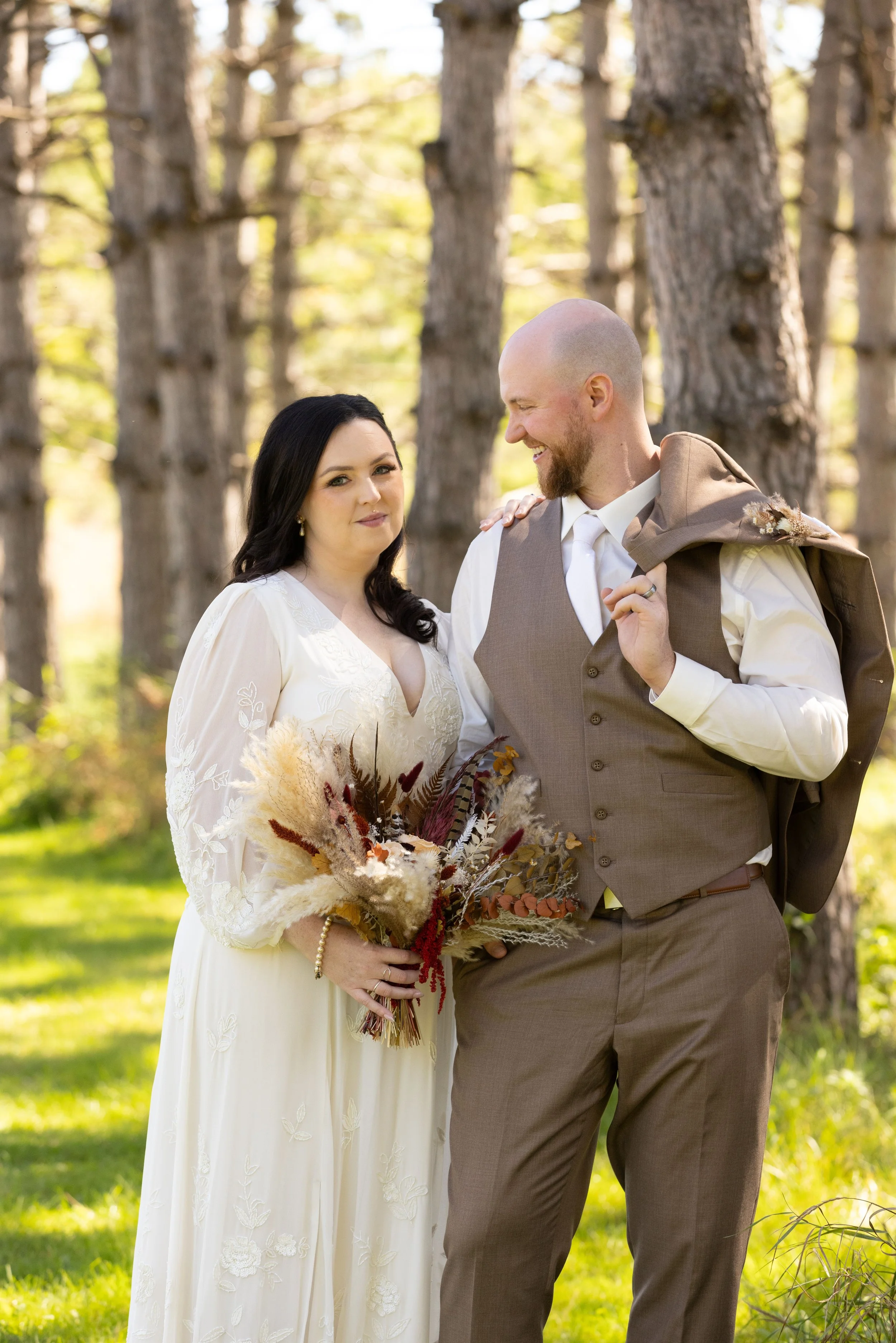 A couple in wedding attire standing outdoors among trees, with the woman holding a bouquet of dried flowers and the man holding a jacket over his shoulder, smiling at each other.