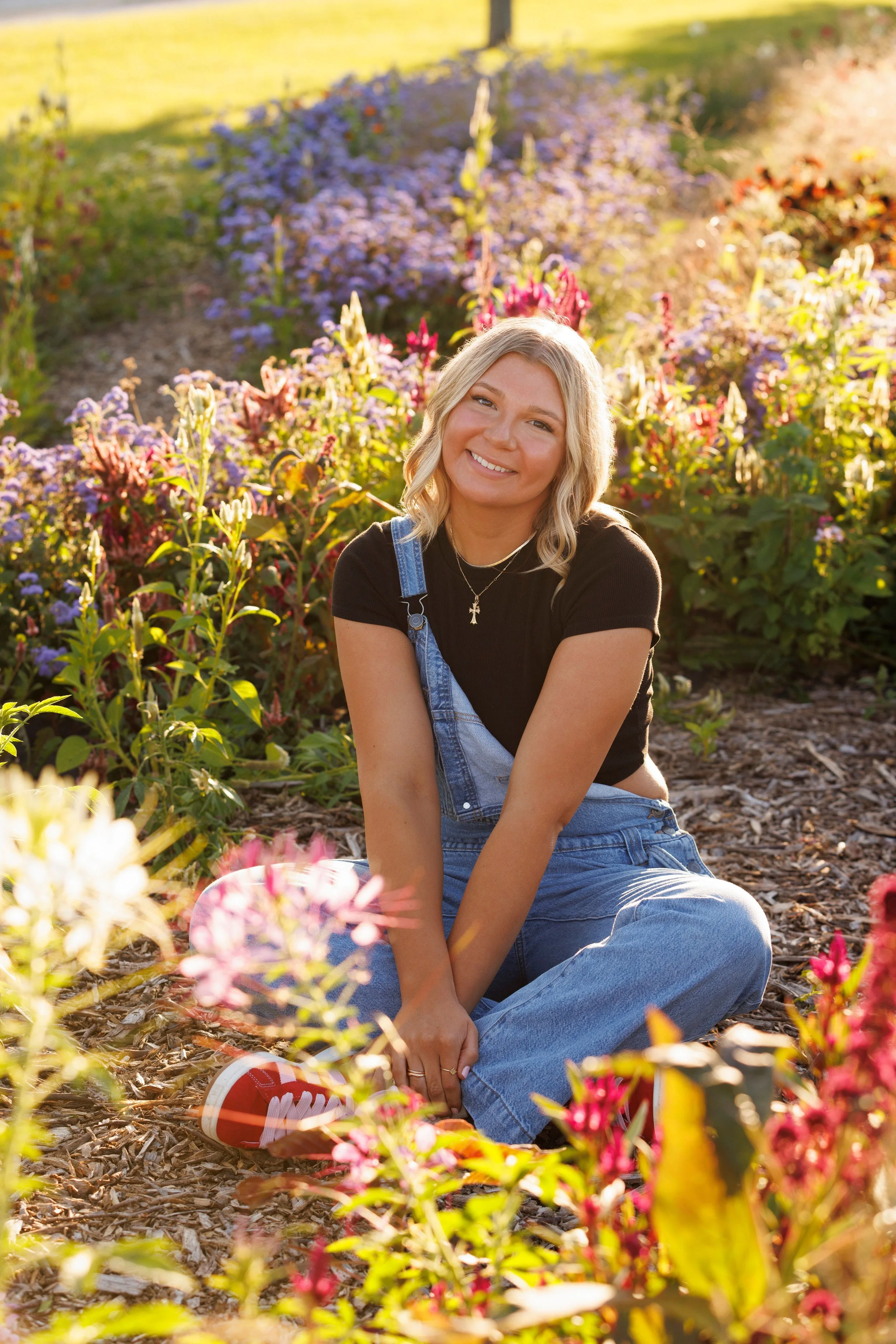 A woman sitting on the ground in a garden with colorful flowers, smiling at the camera during sunset.