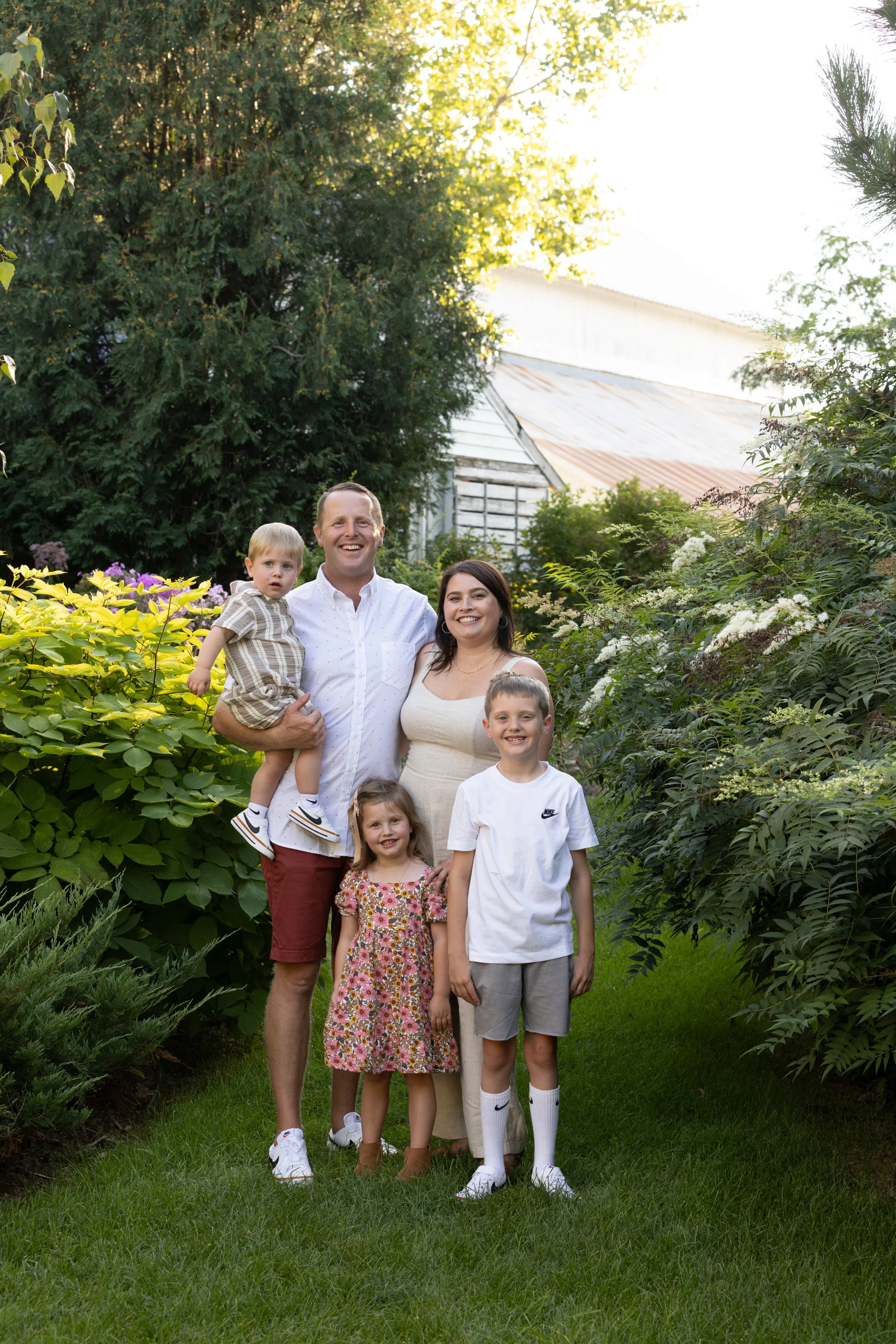 A family of five stands in a lush garden with trees and flowering bushes, smiling at the camera on a sunny day.
