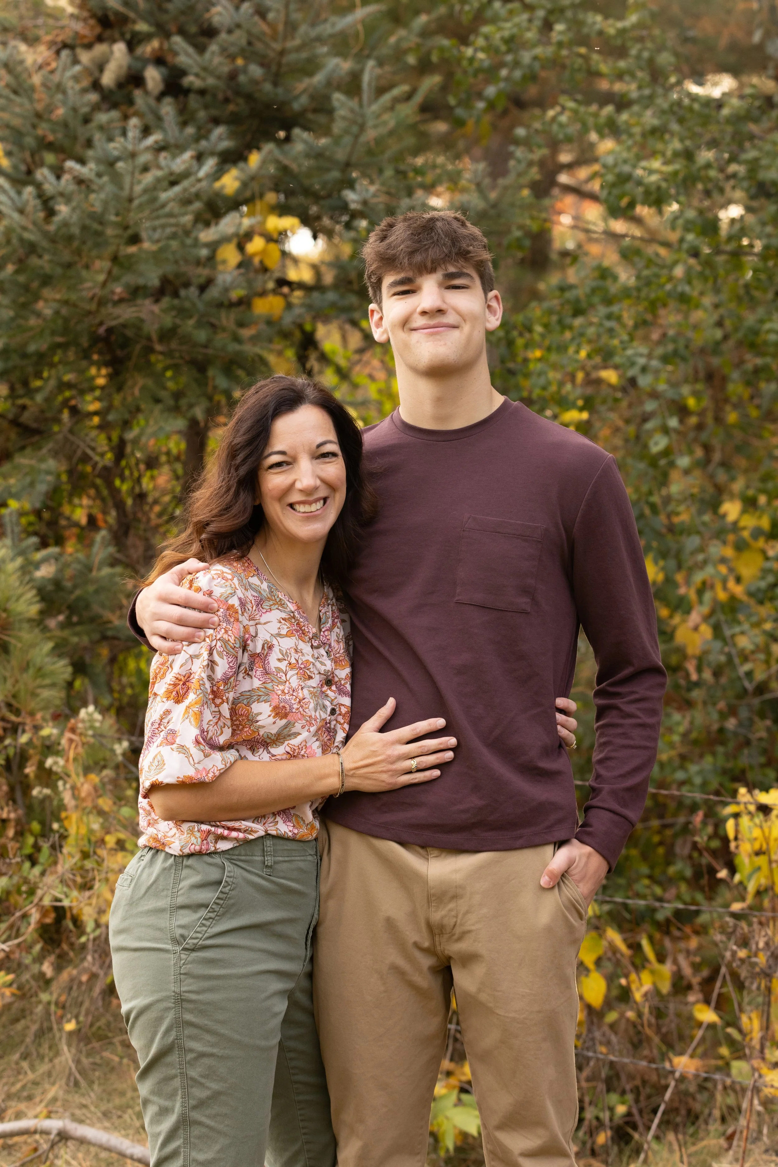 A woman and a teenage boy smiling and embracing outdoors in front of autumn foliage.