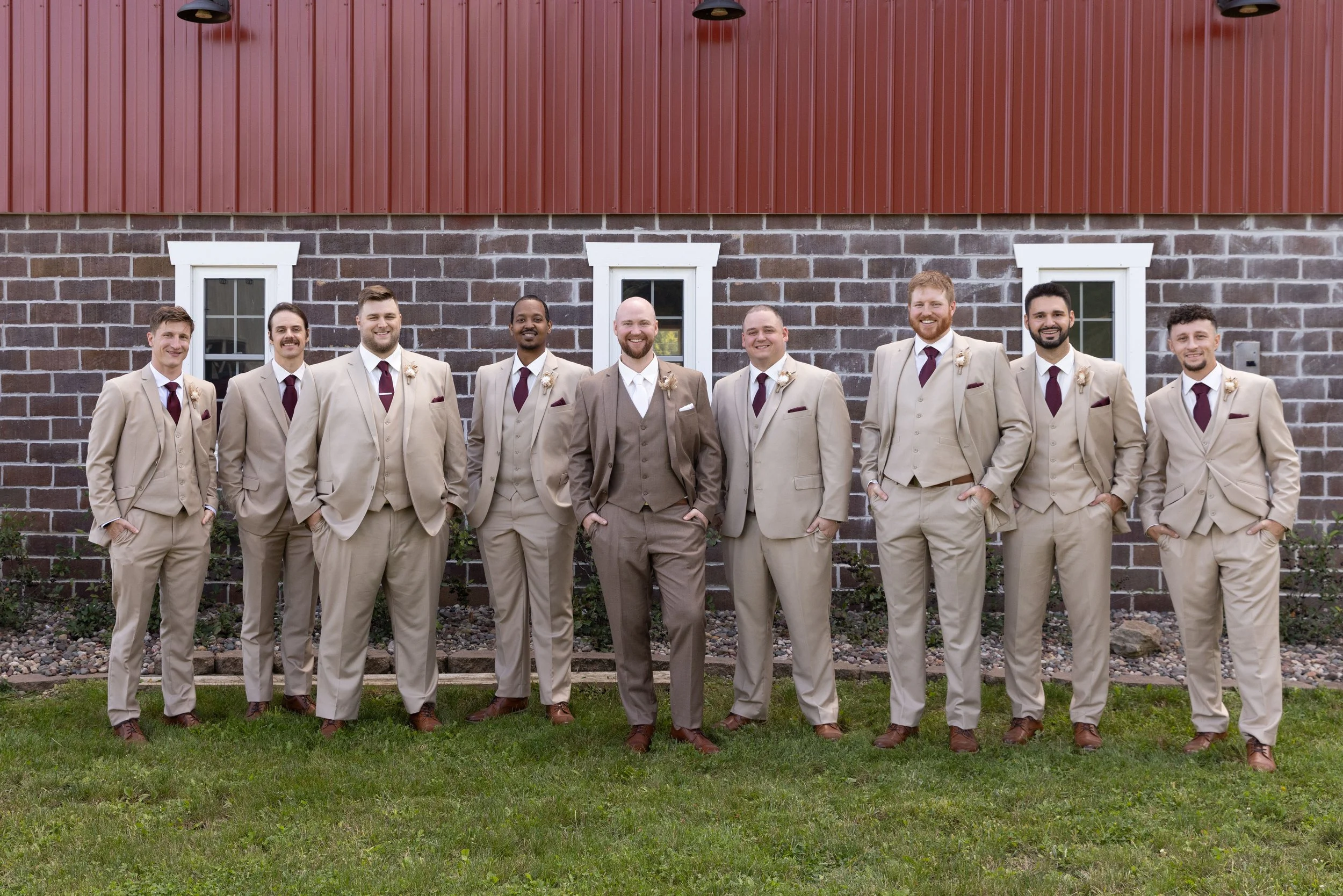 A group of nine men in beige suits, white shirts, and maroon ties posing outdoors in front of a brick building.