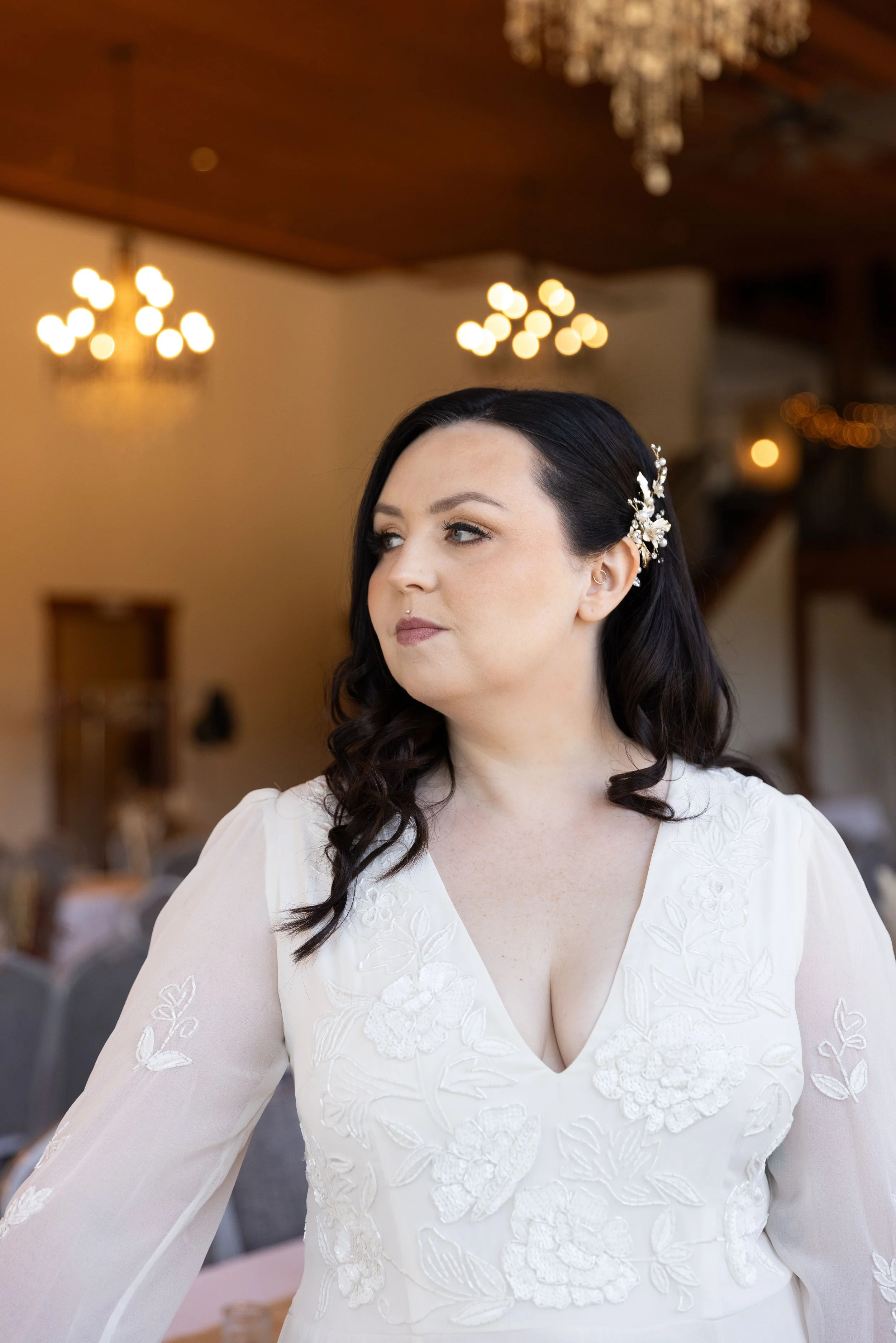 A woman with dark hair in loose curls, wearing a white embroidered dress with a deep neckline and floral embellishments, standing indoors with warm lighting and chandeliers in the background.