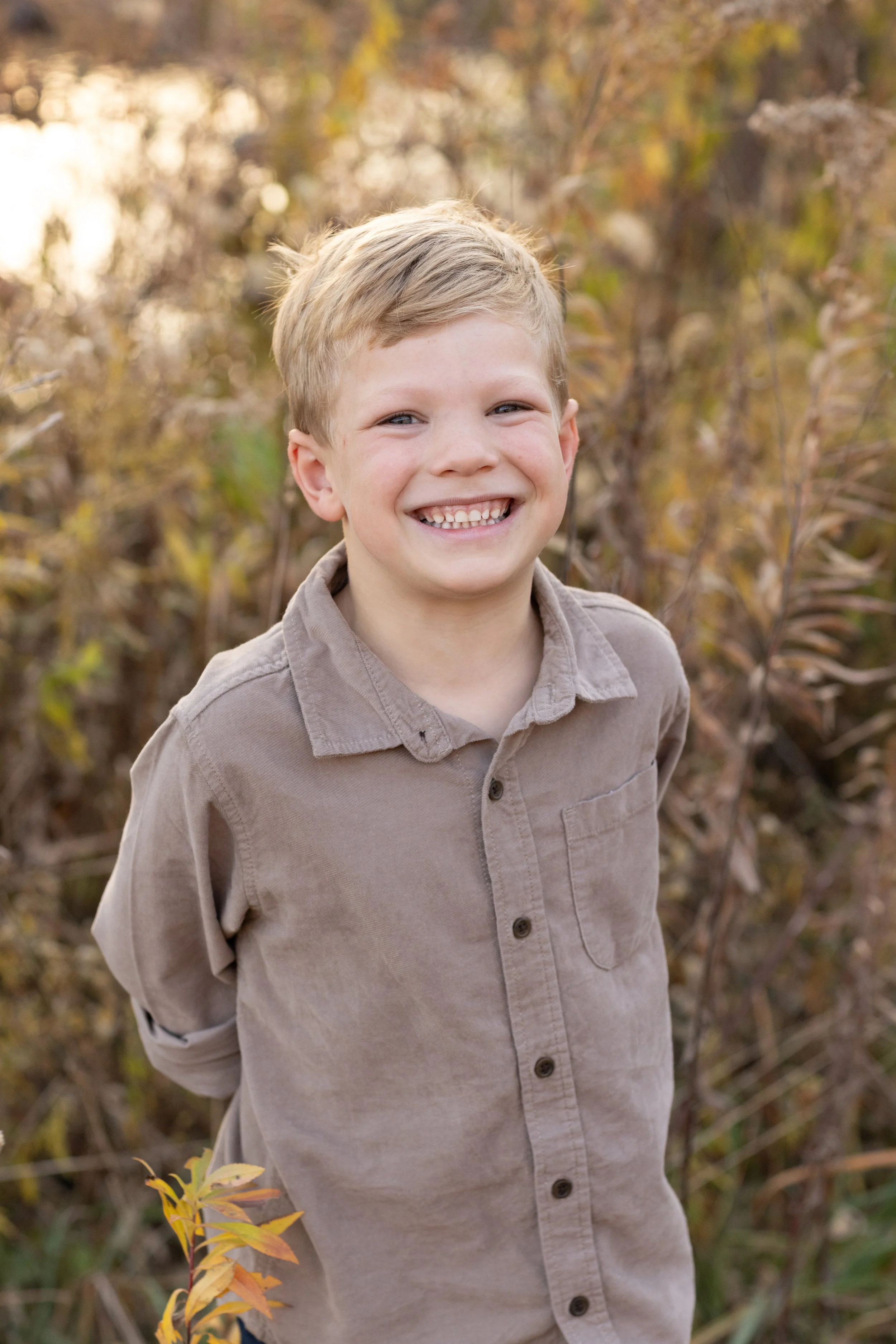 A smiling young boy with short blonde hair, wearing a beige button-up shirt, standing outdoors in a field of tall, dried plants and trees with autumn-colored leaves in the background.