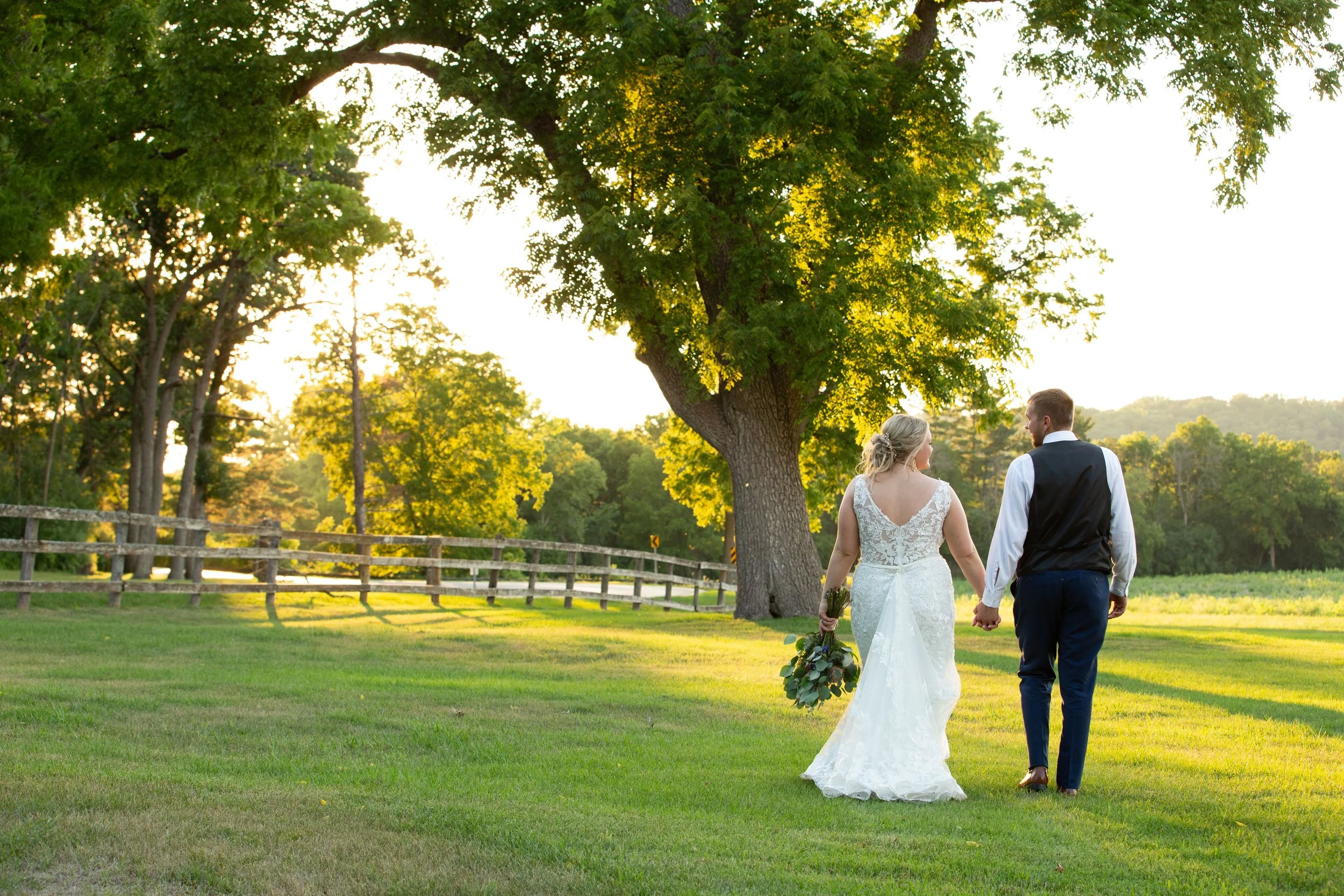 A bride and groom walking hand in hand on a grassy field with large trees and a wooden fence in the background during sunset.