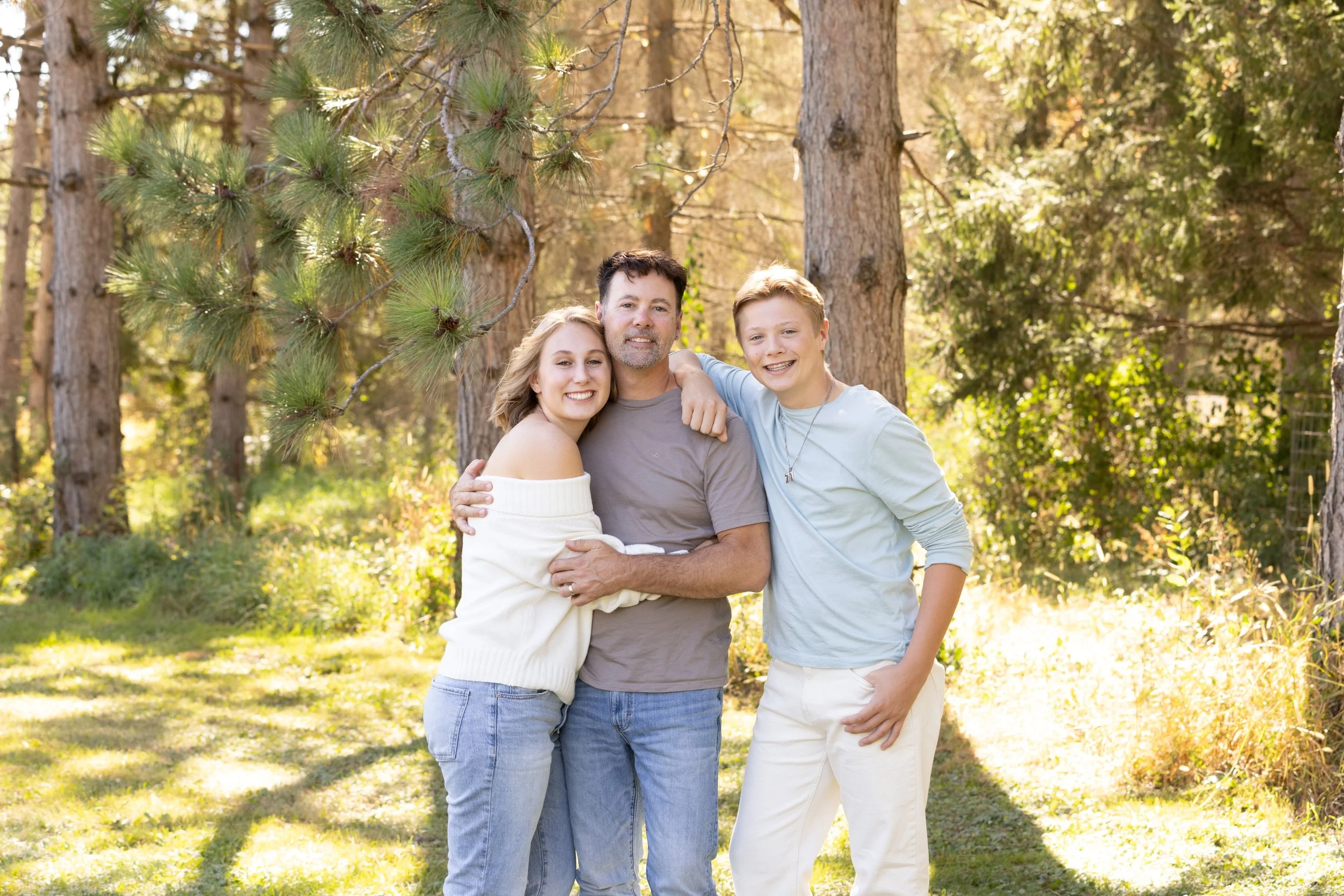 A family of three embracing outdoors in a wooded area on a sunny day, smiling at the camera.