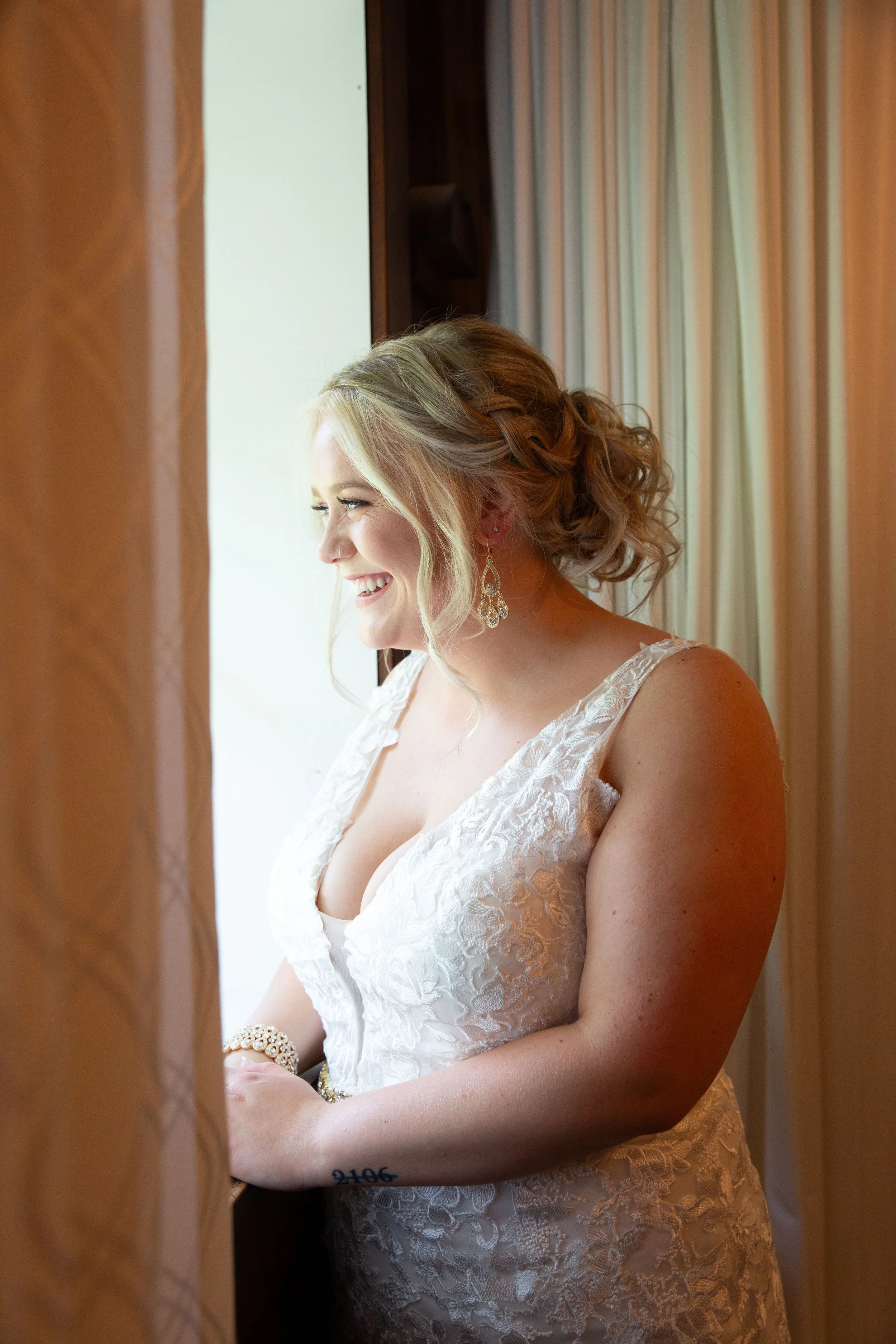 A bride with blonde hair styled in loose curls, wearing a white lace wedding dress, earrings, and a bracelet, smiling as she looks out a window.