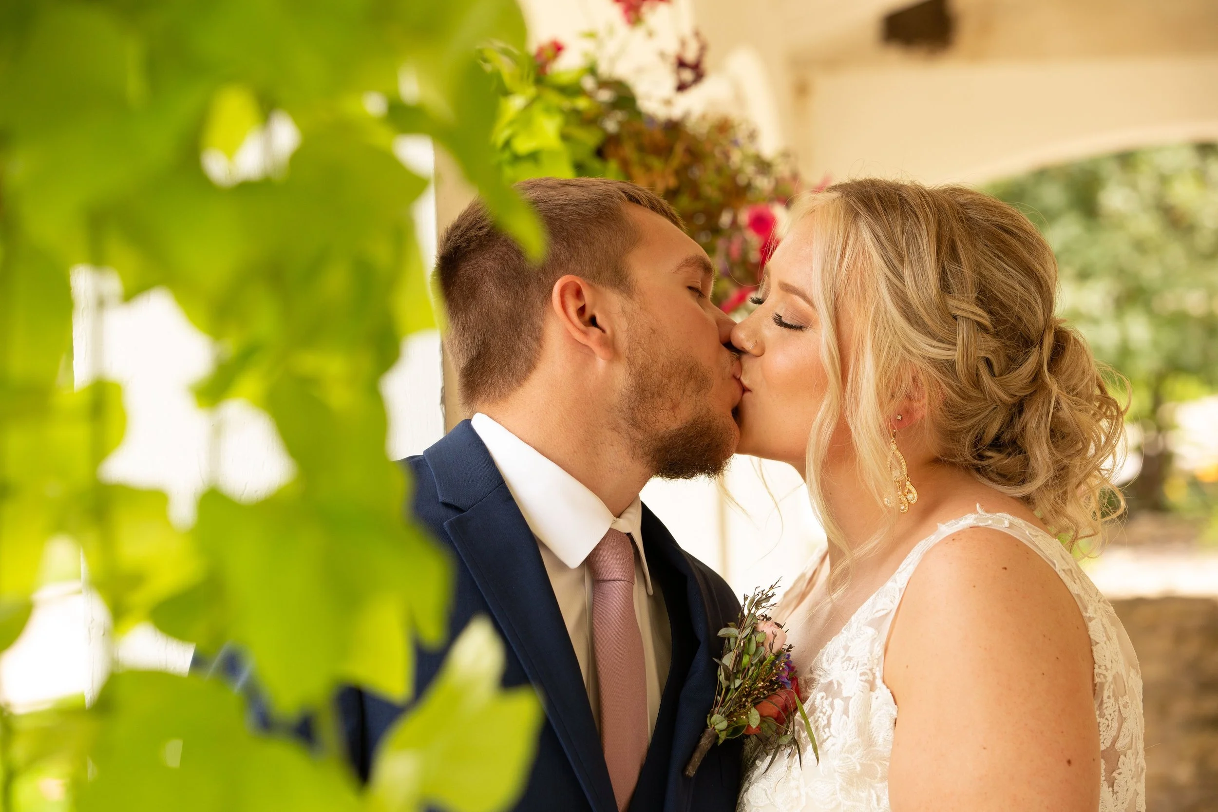 A couple is sharing a kiss at their wedding, with greenery and flowers in the background.
