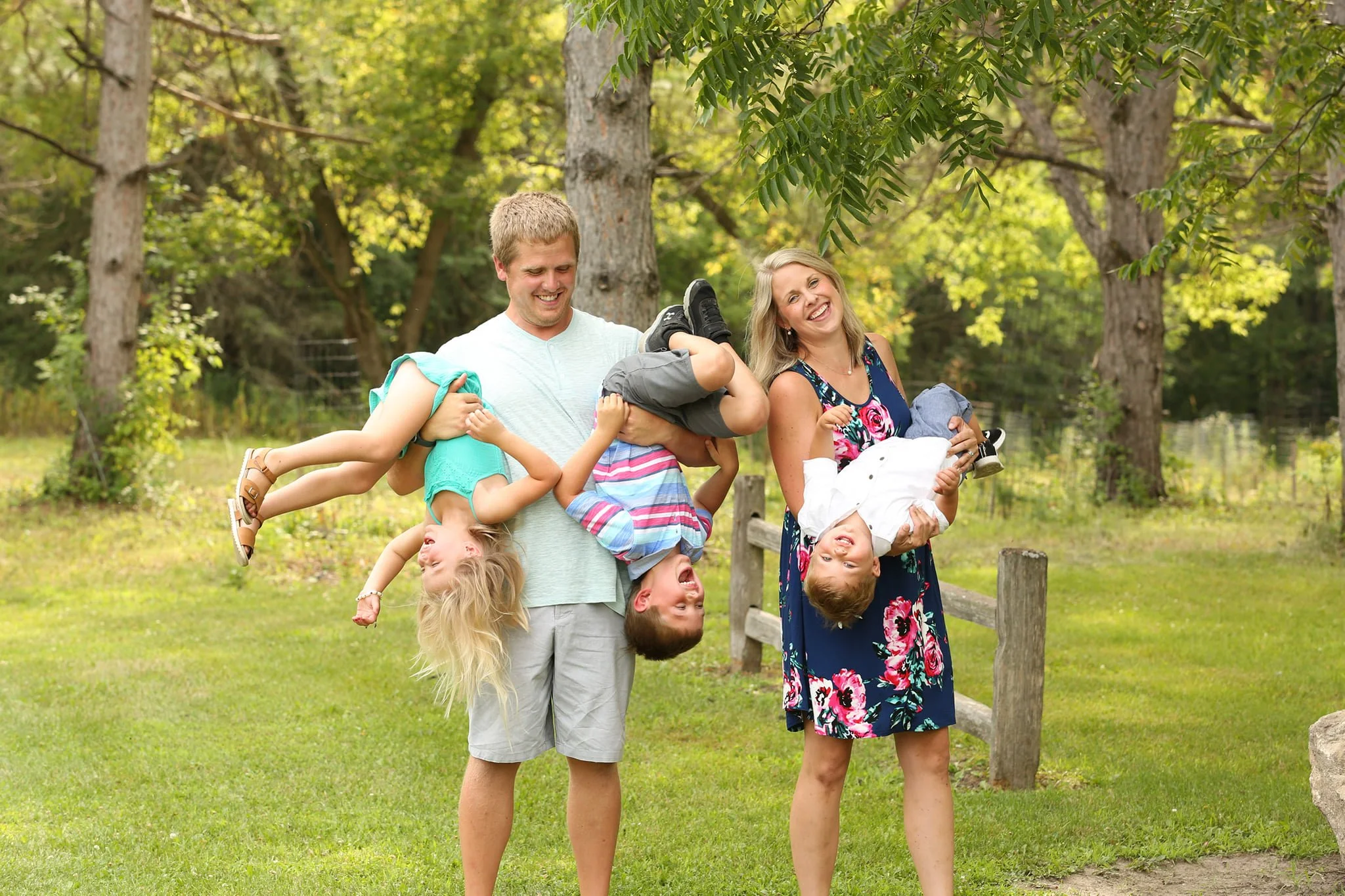 A family of four playing in a park, with children hanging upside down from their parents' arms, all smiling and laughing, with trees and grass in the background.