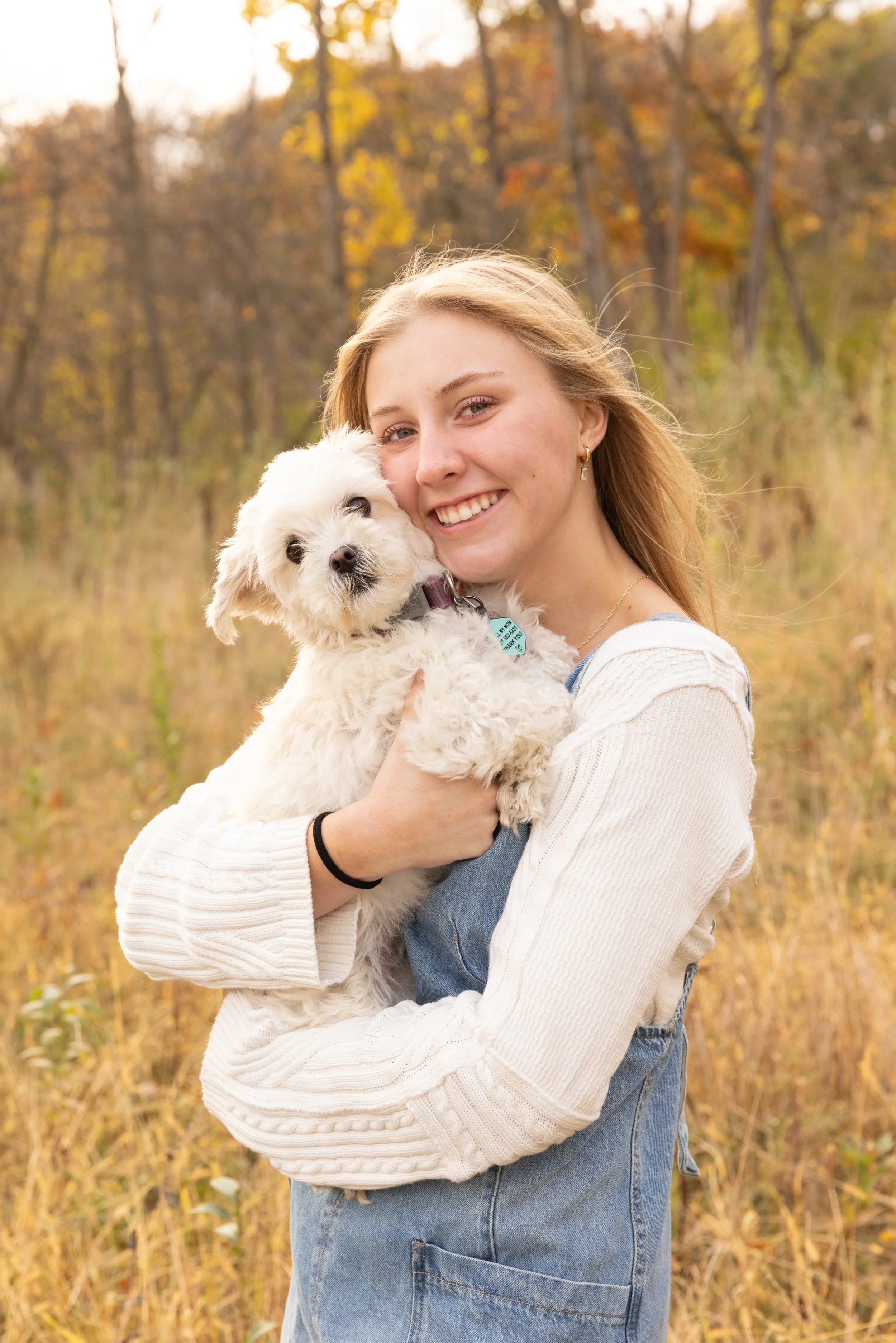 A young woman holding a small fluffy white dog outdoors in a field during autumn with fall foliage in the background.