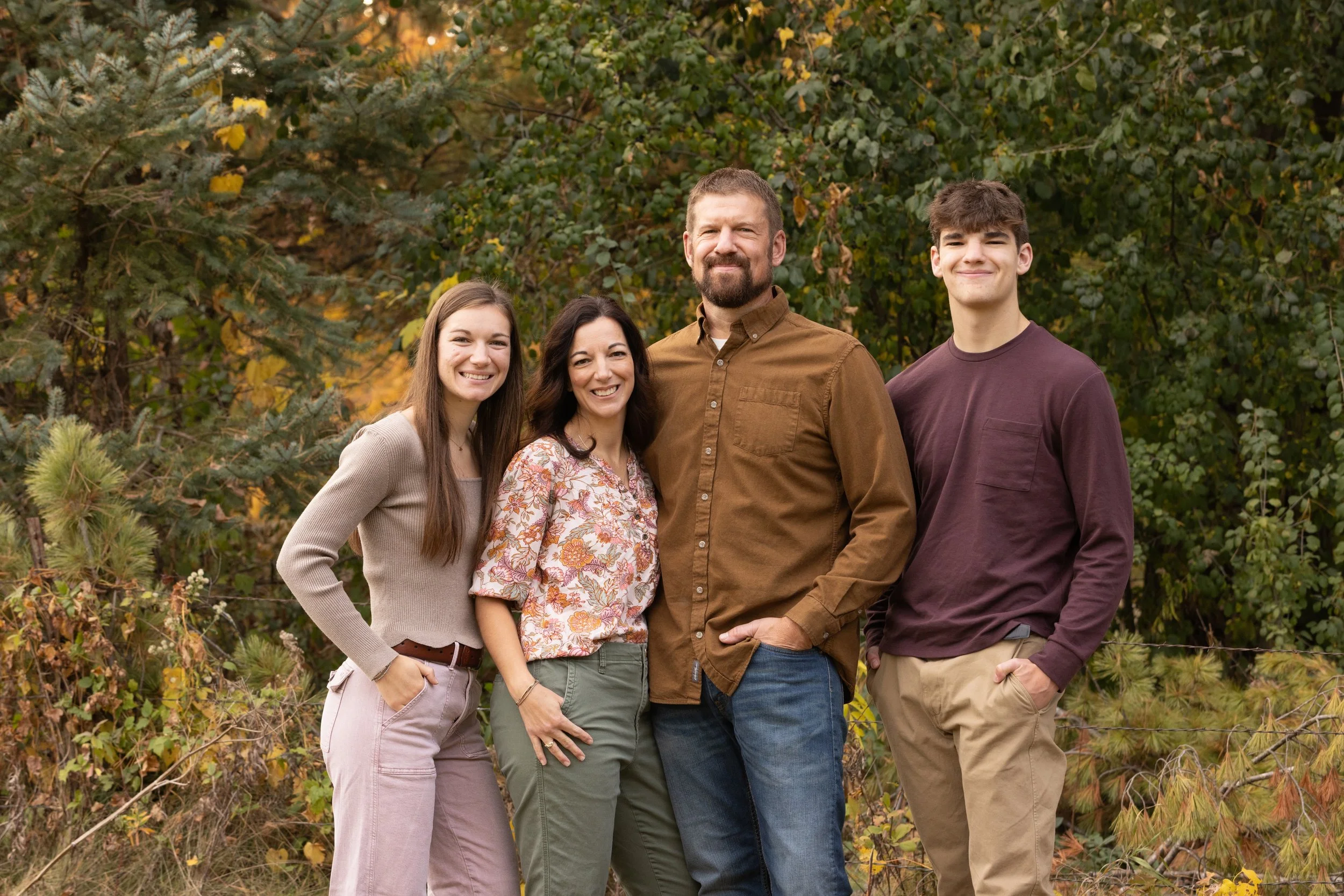 A family of four standing outdoors in a forested area during autumn, smiling at the camera.