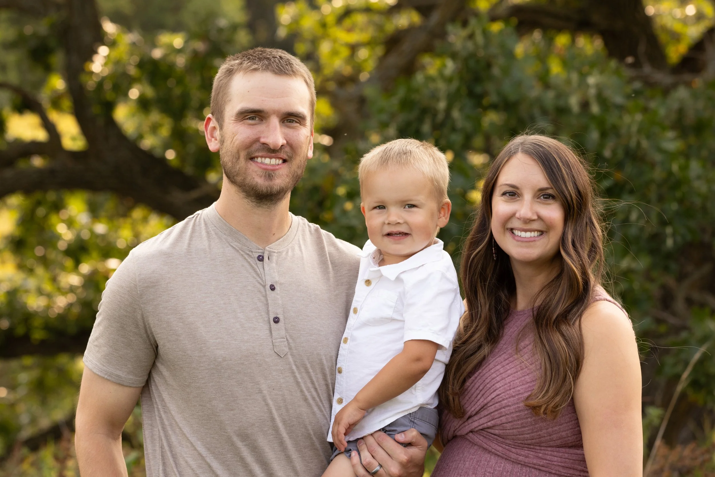 A family of three outdoors, consisting of a man, woman, and young boy, smiling at the camera with trees in the background.