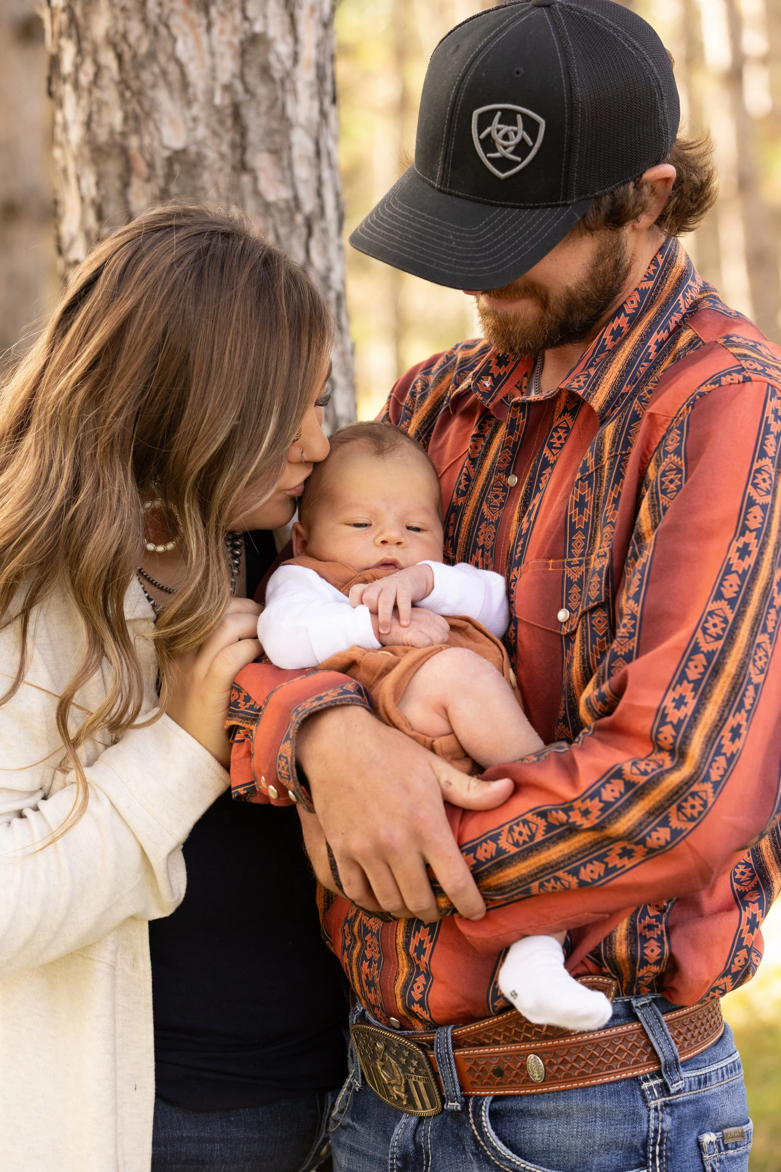A young couple holding their baby outdoors in a forest with trees in the background.