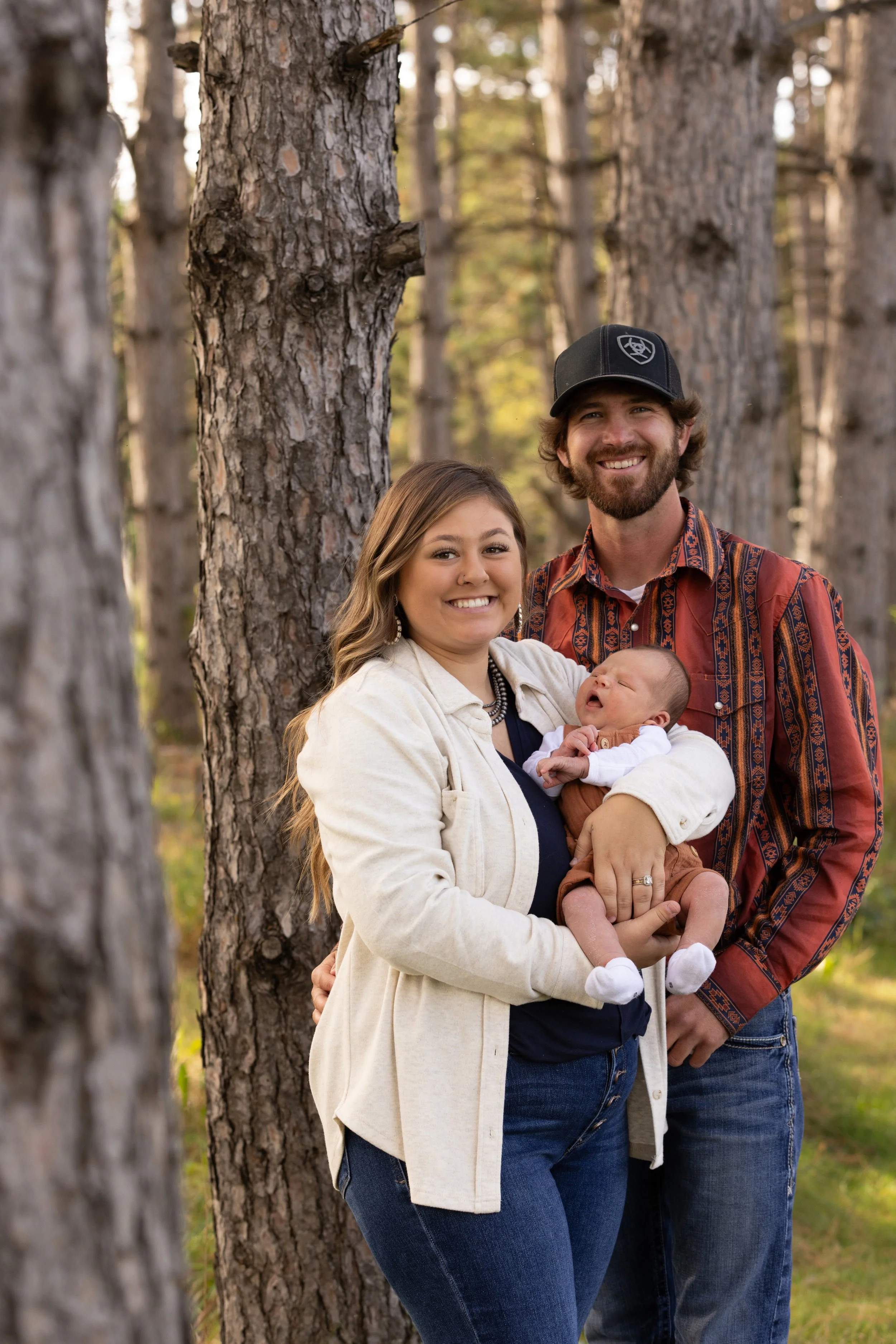 A happy family of three standing in a forest with tall trees during daytime. The woman holds a newborn baby, and the man stands beside her, smiling.