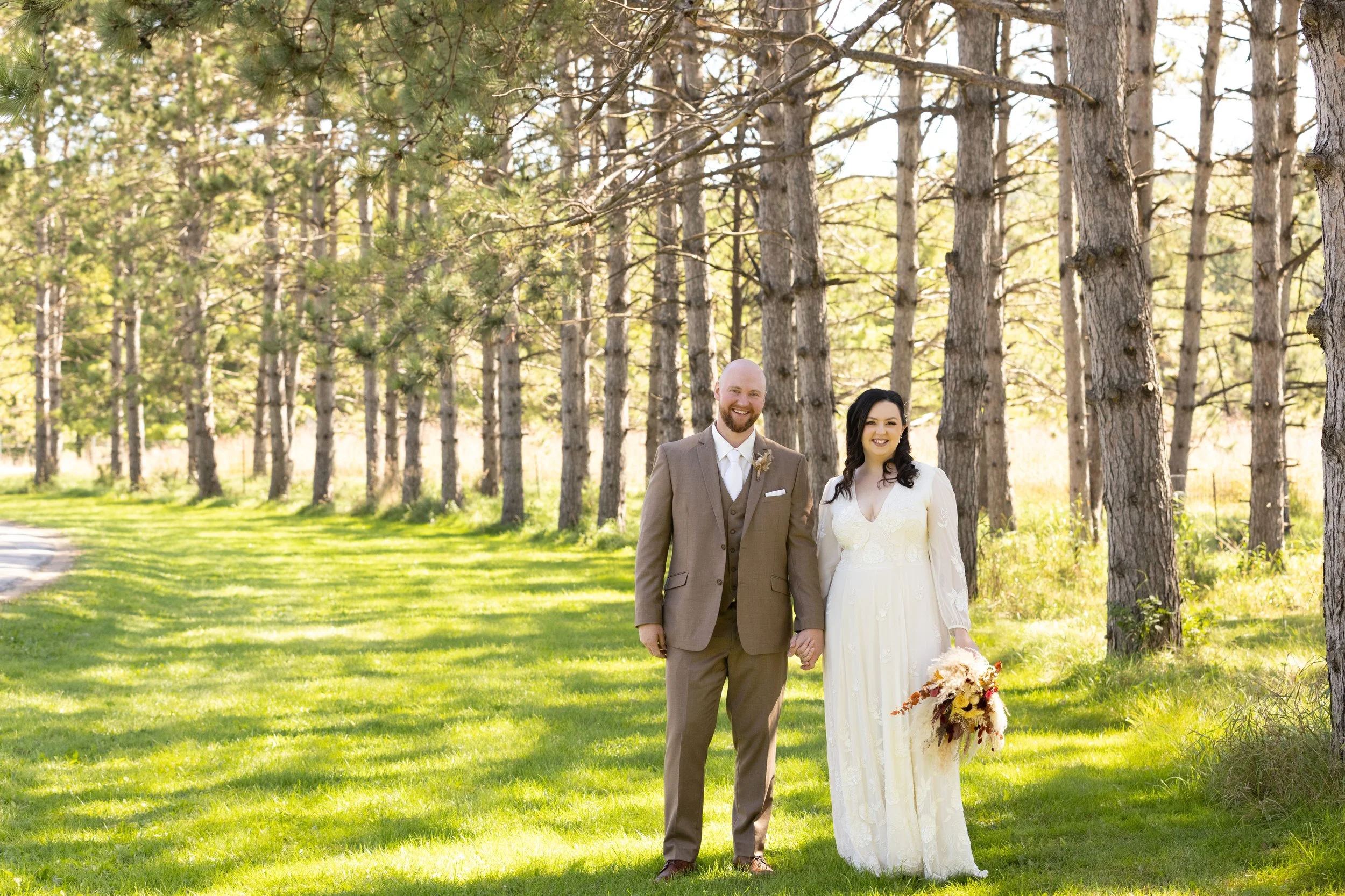 Couple in wedding attire holding hands in a forested outdoor setting with tall trees and green grass.