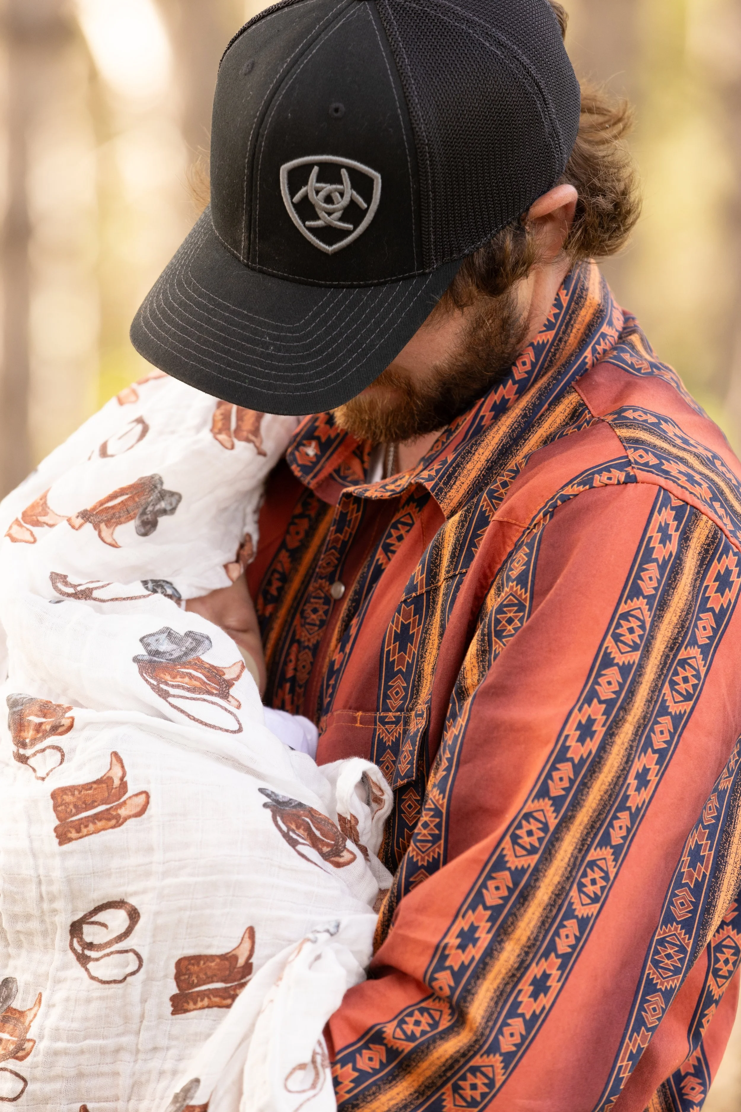 A man with a beard wearing a black cap and a patterned orange and blue shirt is holding a baby wrapped in a white blanket with animal print. The background shows a blurred outdoor setting with trees.