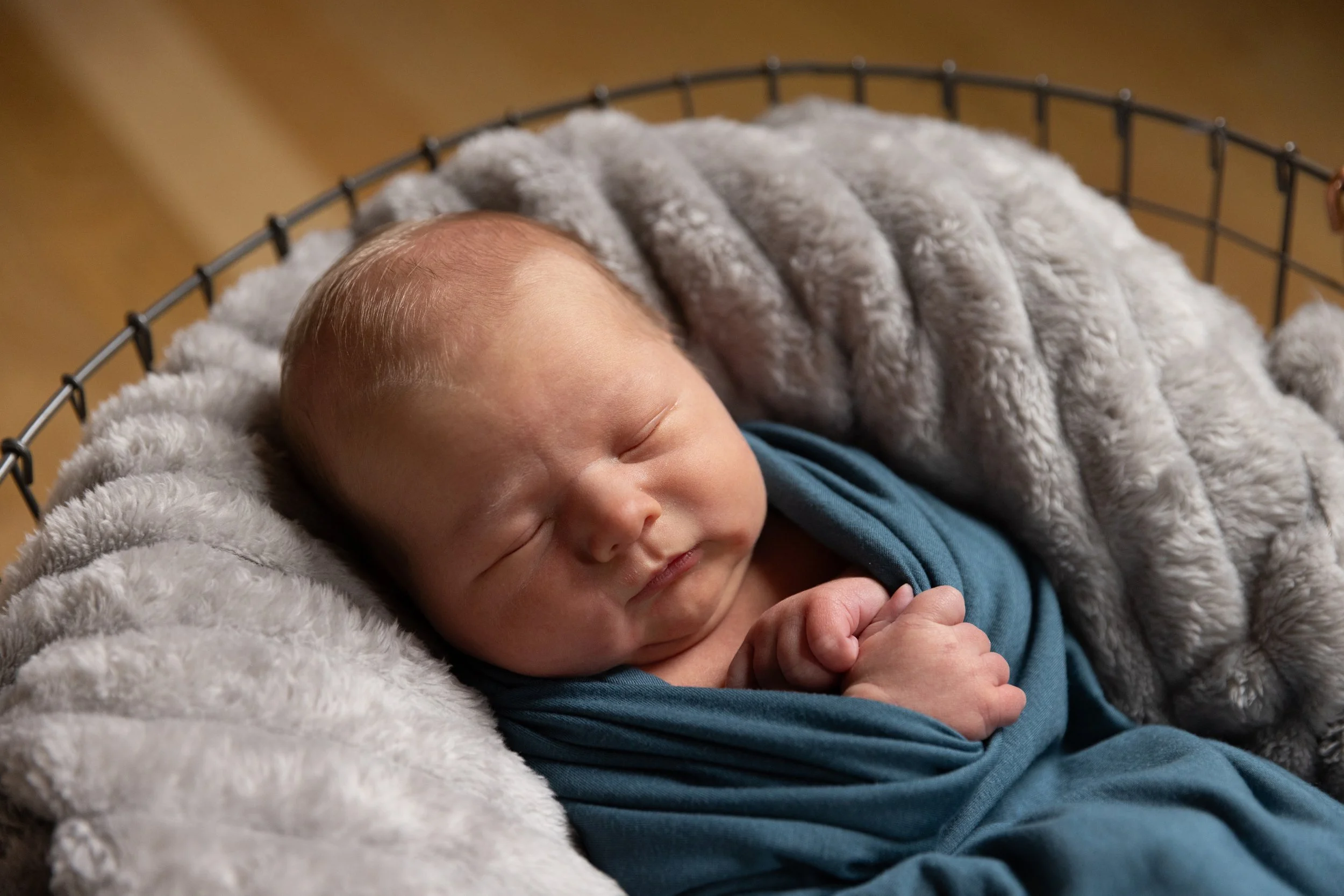 A sleeping newborn baby wrapped in a blue swaddle, resting on a soft gray blanket in a wire basket.