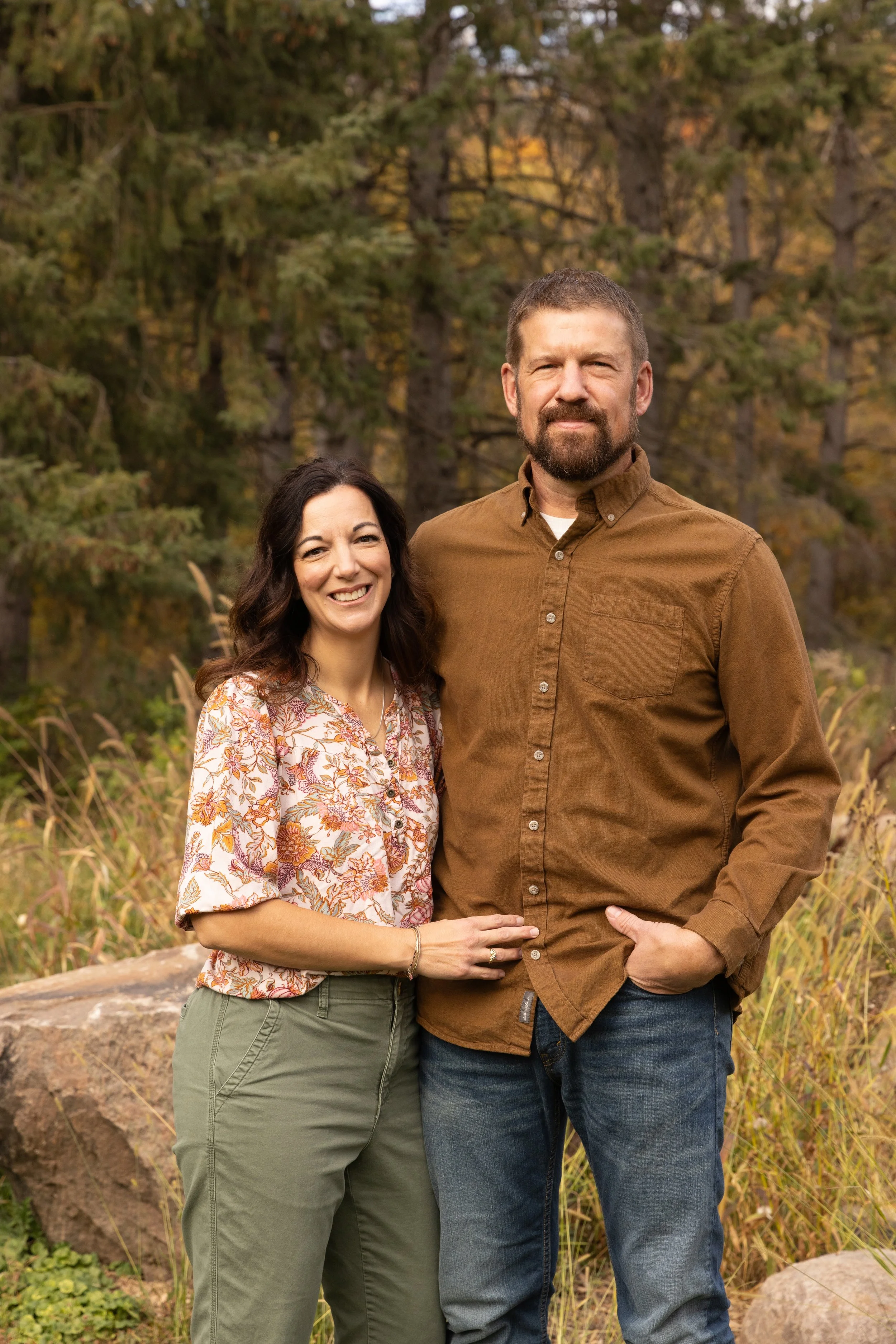 A smiling woman and a man stand outdoors in front of trees and fall foliage, with the woman placing her hand on the man's waist.