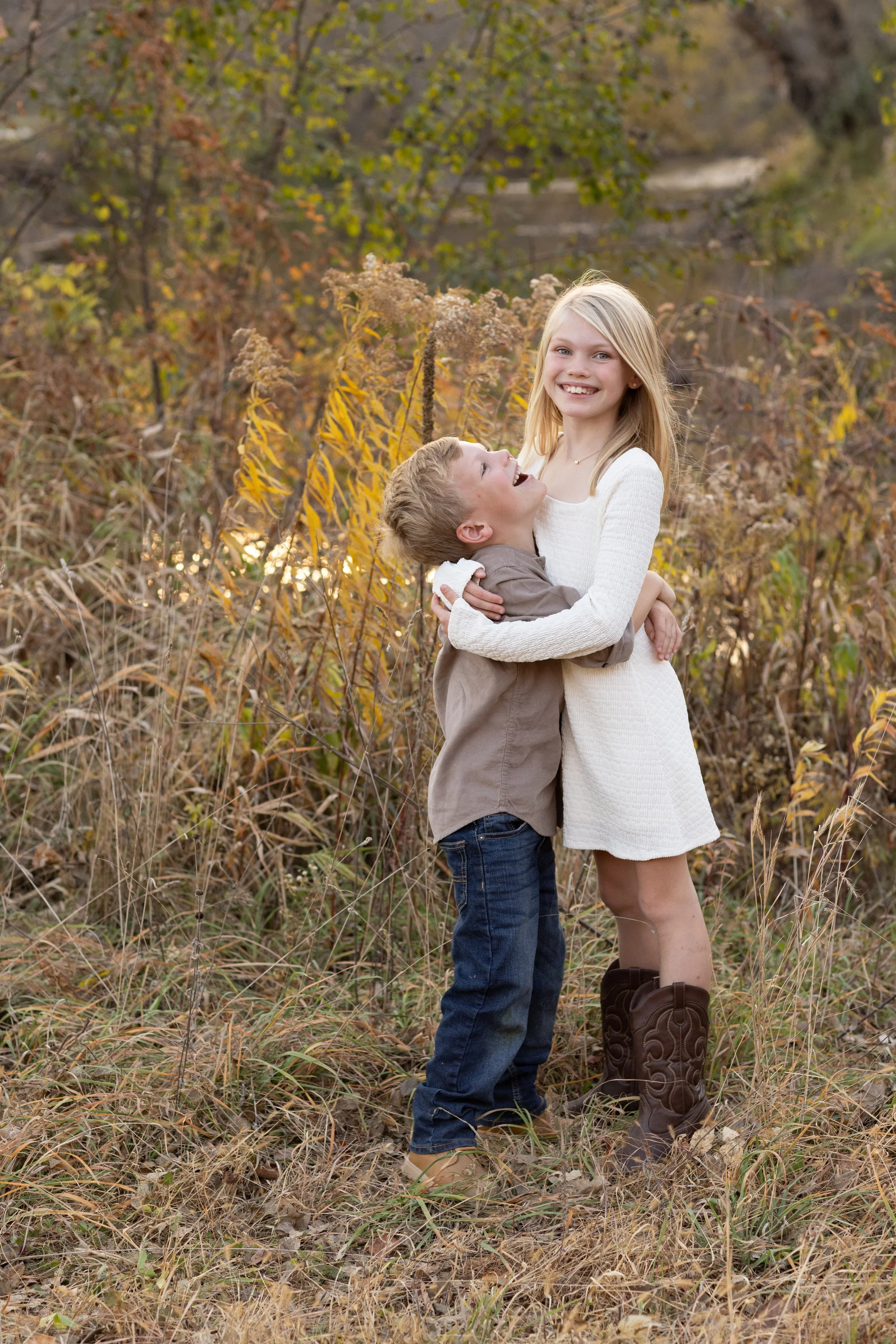 A young girl and a boy embrace in a field of autumn plants and trees, smiling at each other with the girl holding the boy. The girl wears a white dress and brown cowboy boots, and the boy wears a gray shirt and jeans.
