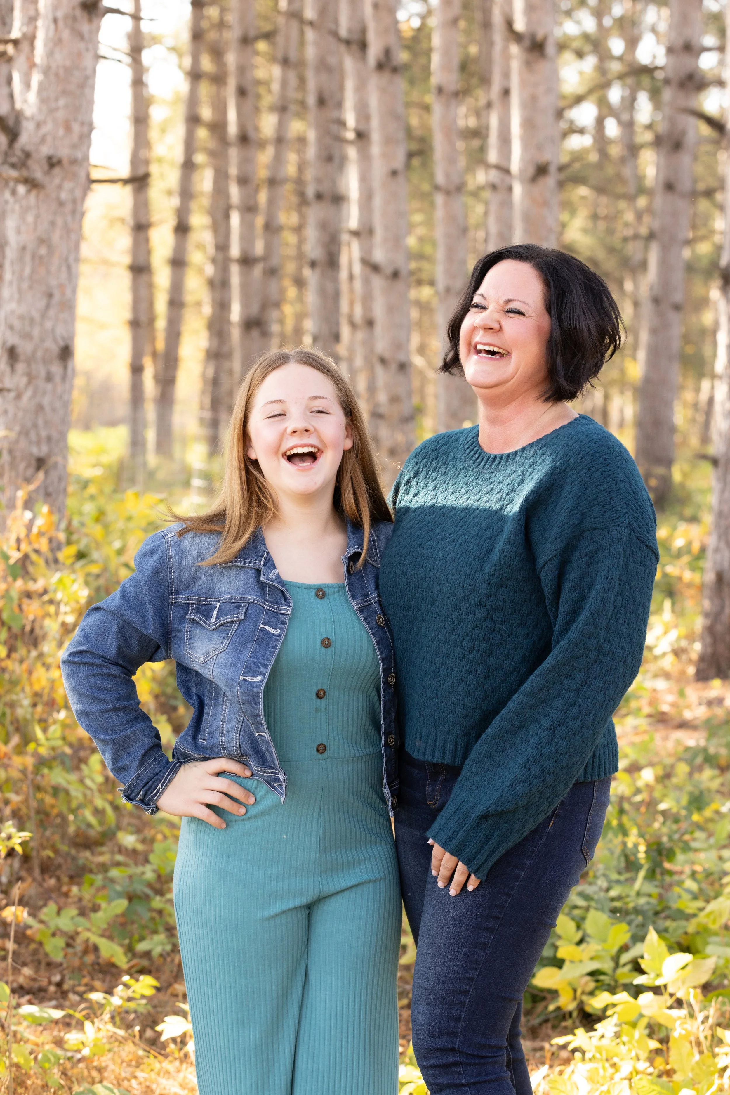 Woman and girl laughing outdoors in a forest with trees and autumn foliage.