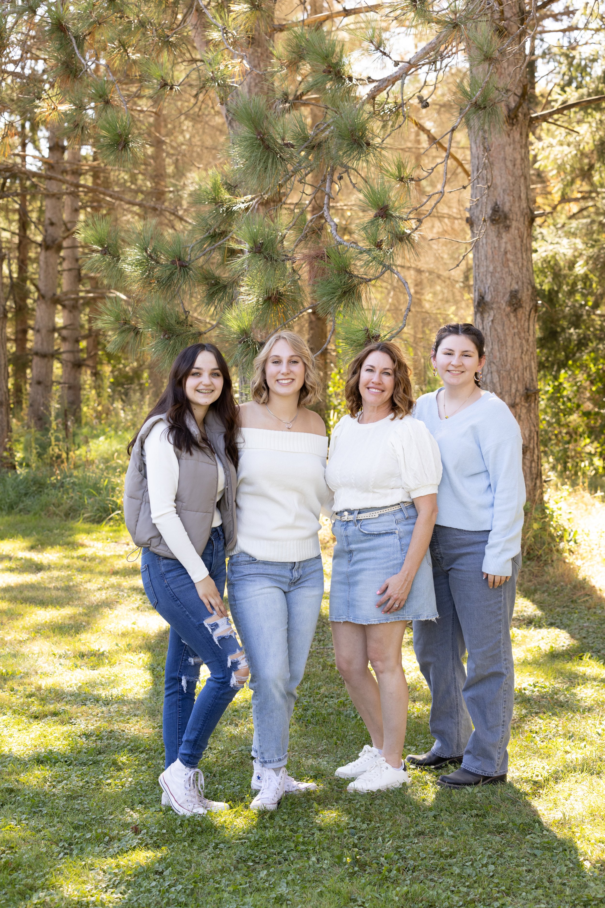 Four women standing outdoors in a forest clearing, smiling at the camera, with tall trees and green grass around them.