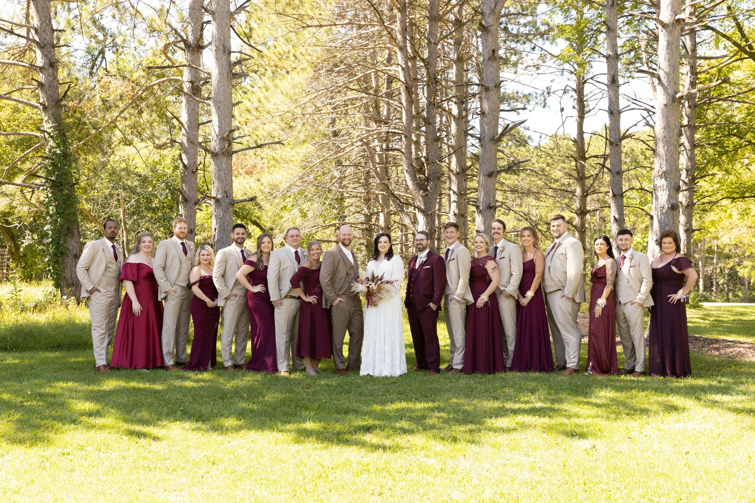A group of people dressed in formal attire outdoors in a wooded park during daytime, including a bride in a white wedding gown holding a bouquet, a groom in a beige suit, bridesmaids in burgundy dresses, and groomsmen in beige suits, all standing on 