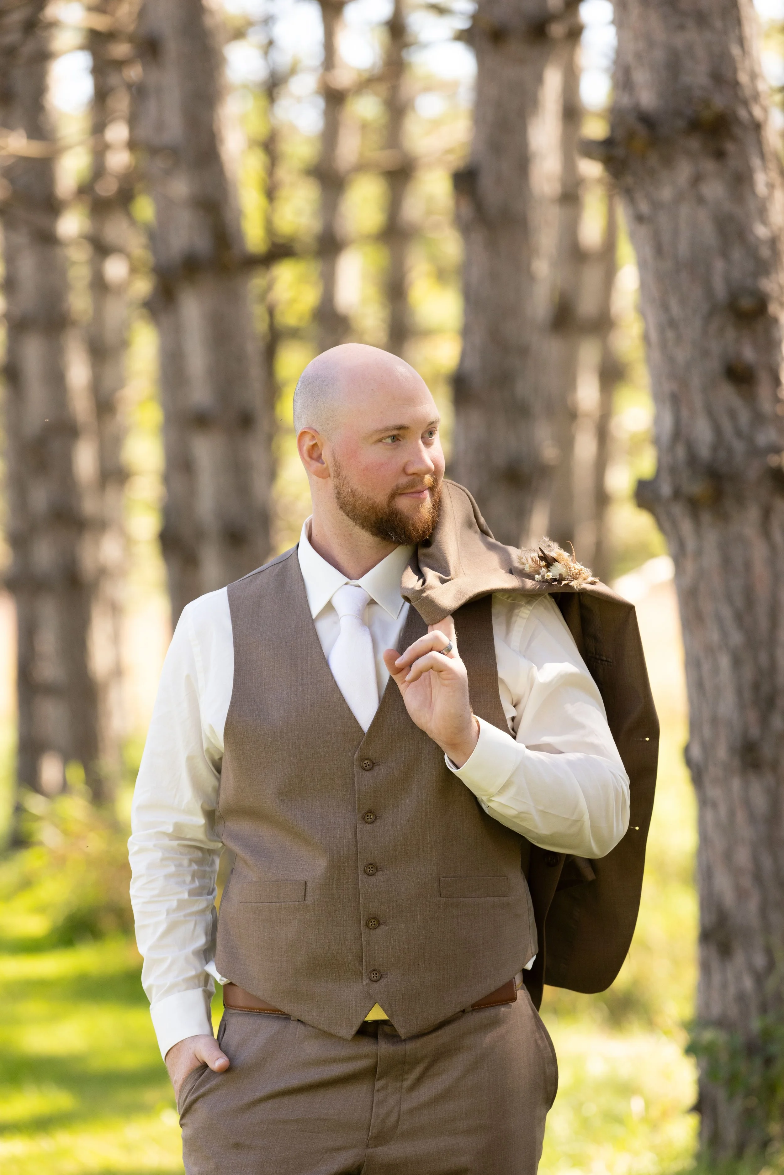 A man in a brown vest, white shirt, and tie, carrying a jacket over his shoulder, standing in a forest with tall trees.