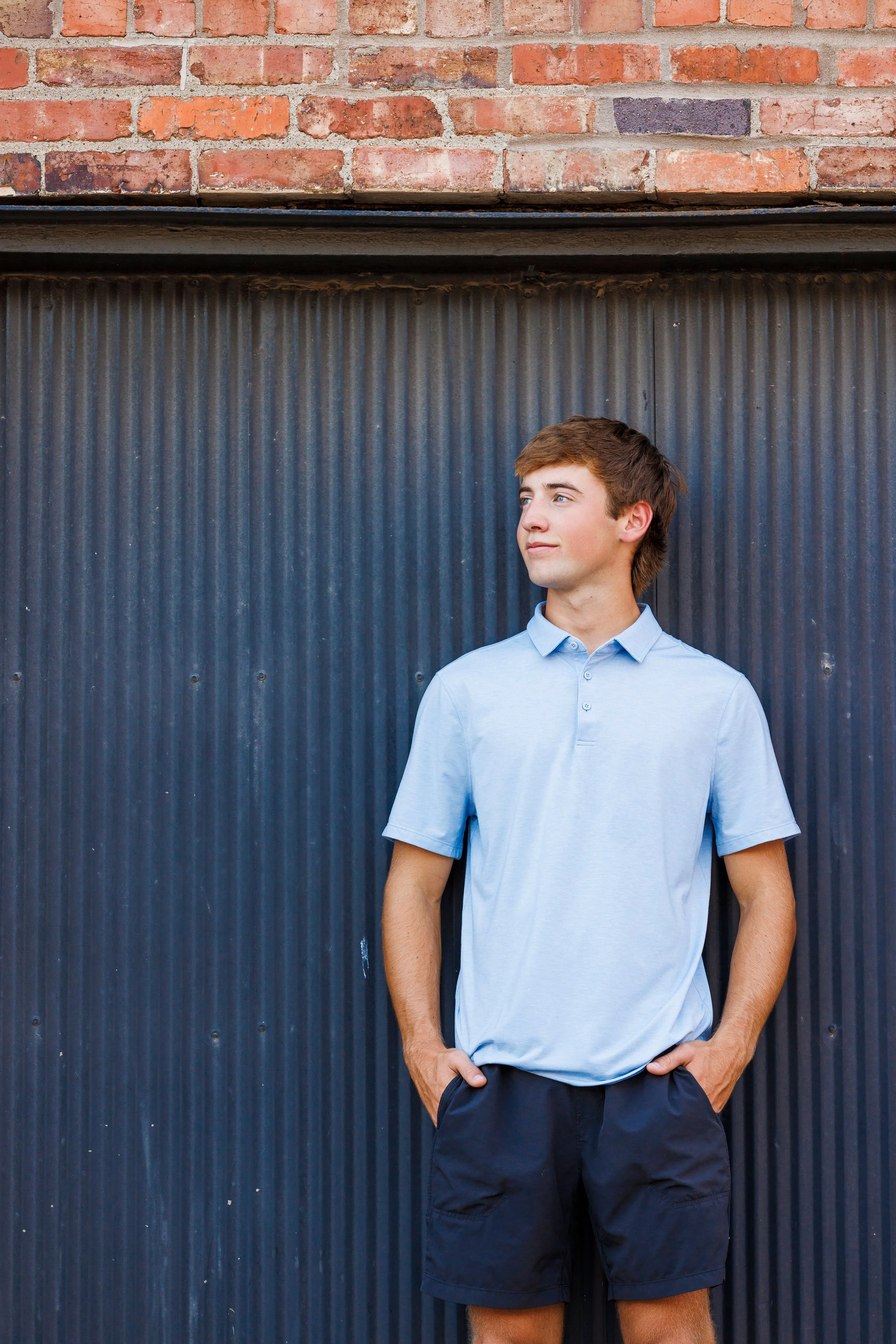 Young man with brown hair, wearing a light blue polo shirt and dark shorts, standing with hands in pockets against a black corrugated metal wall and red brick wall above.