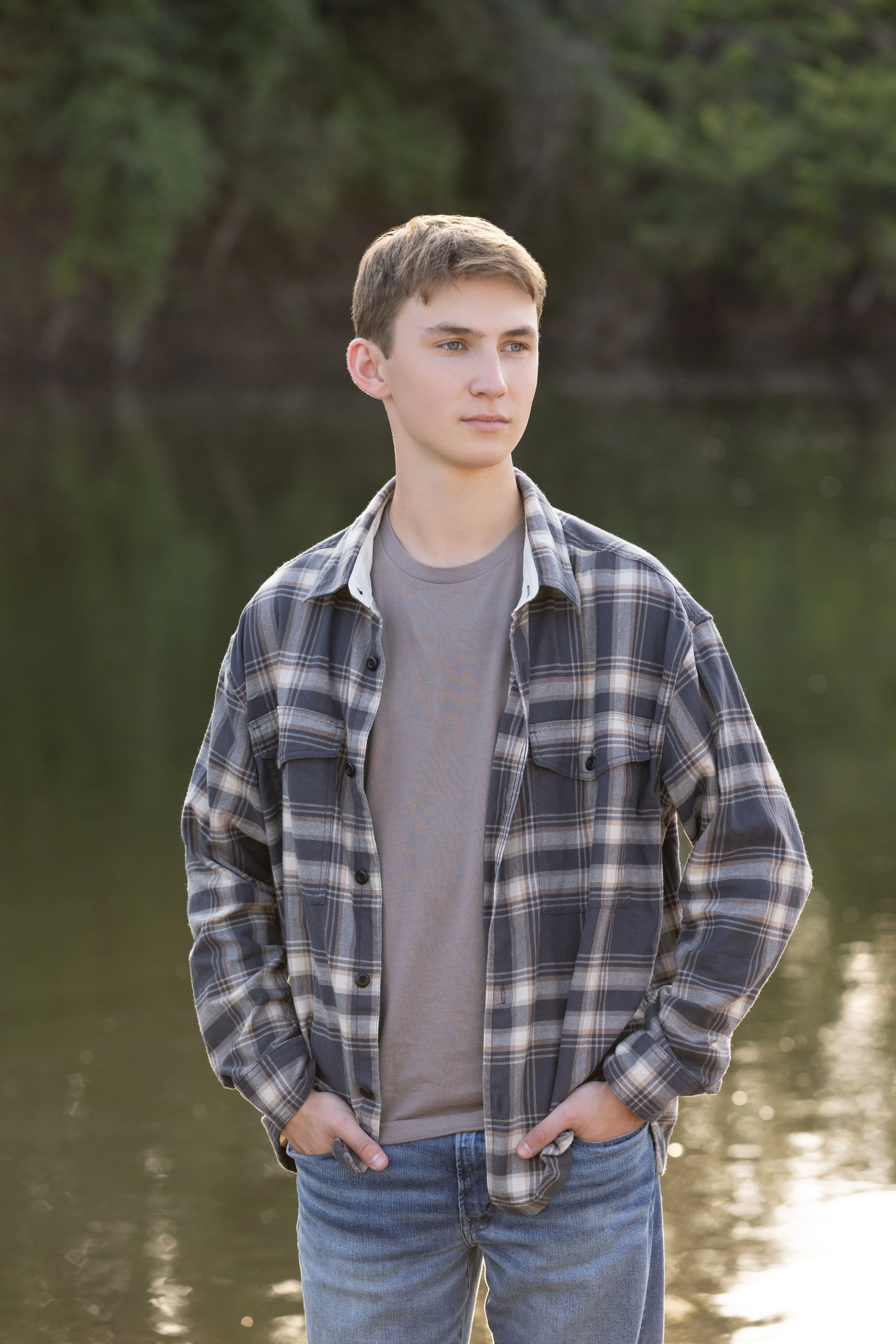 A young man standing outdoors near a body of water, wearing a gray T-shirt, a plaid shirt, and jeans, with hands in pockets, looking to the side.
